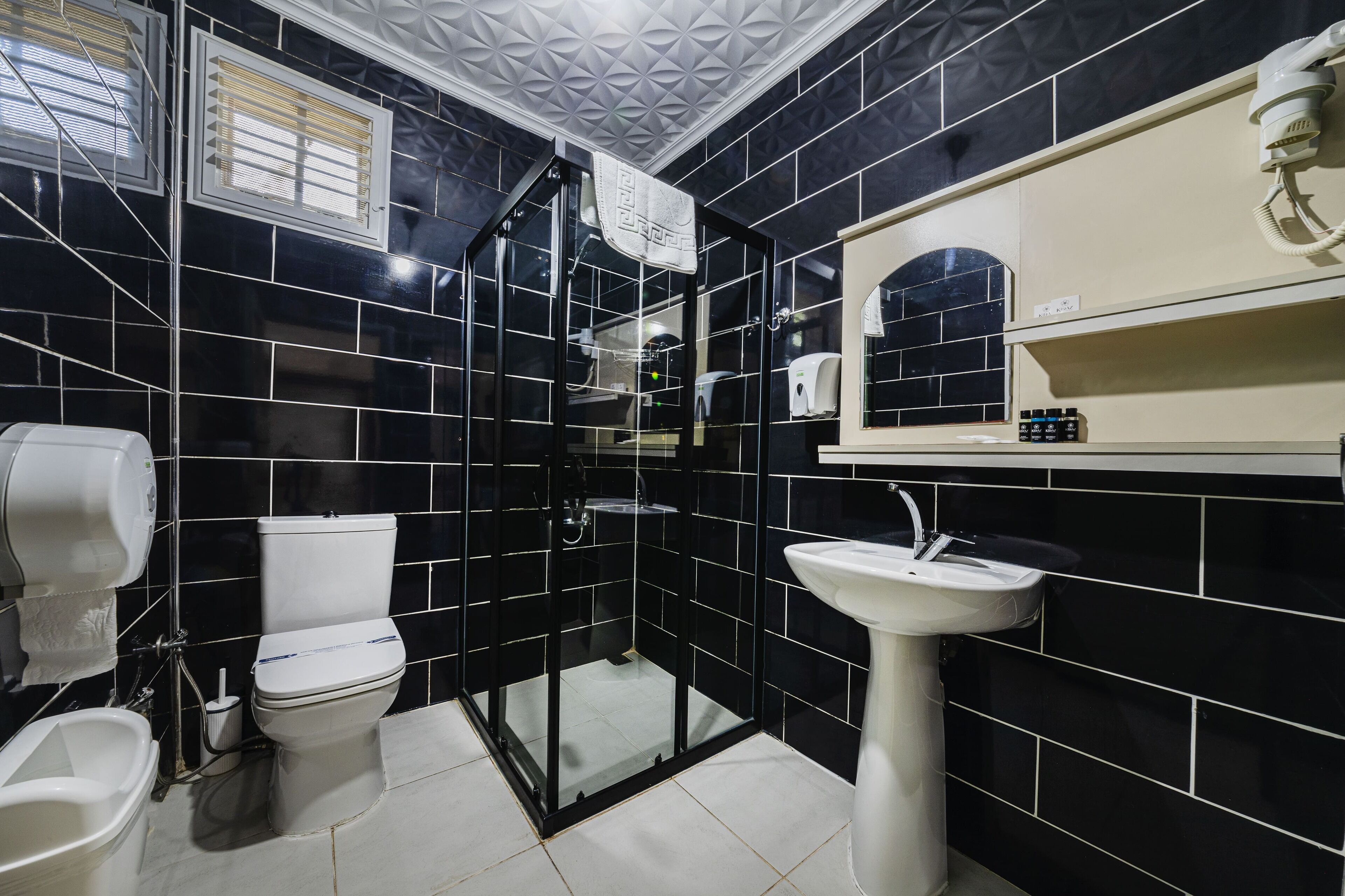 A bathroom with black tiles, a white toilet, a glass-enclosed shower, a white sink, and a towel dispenser.