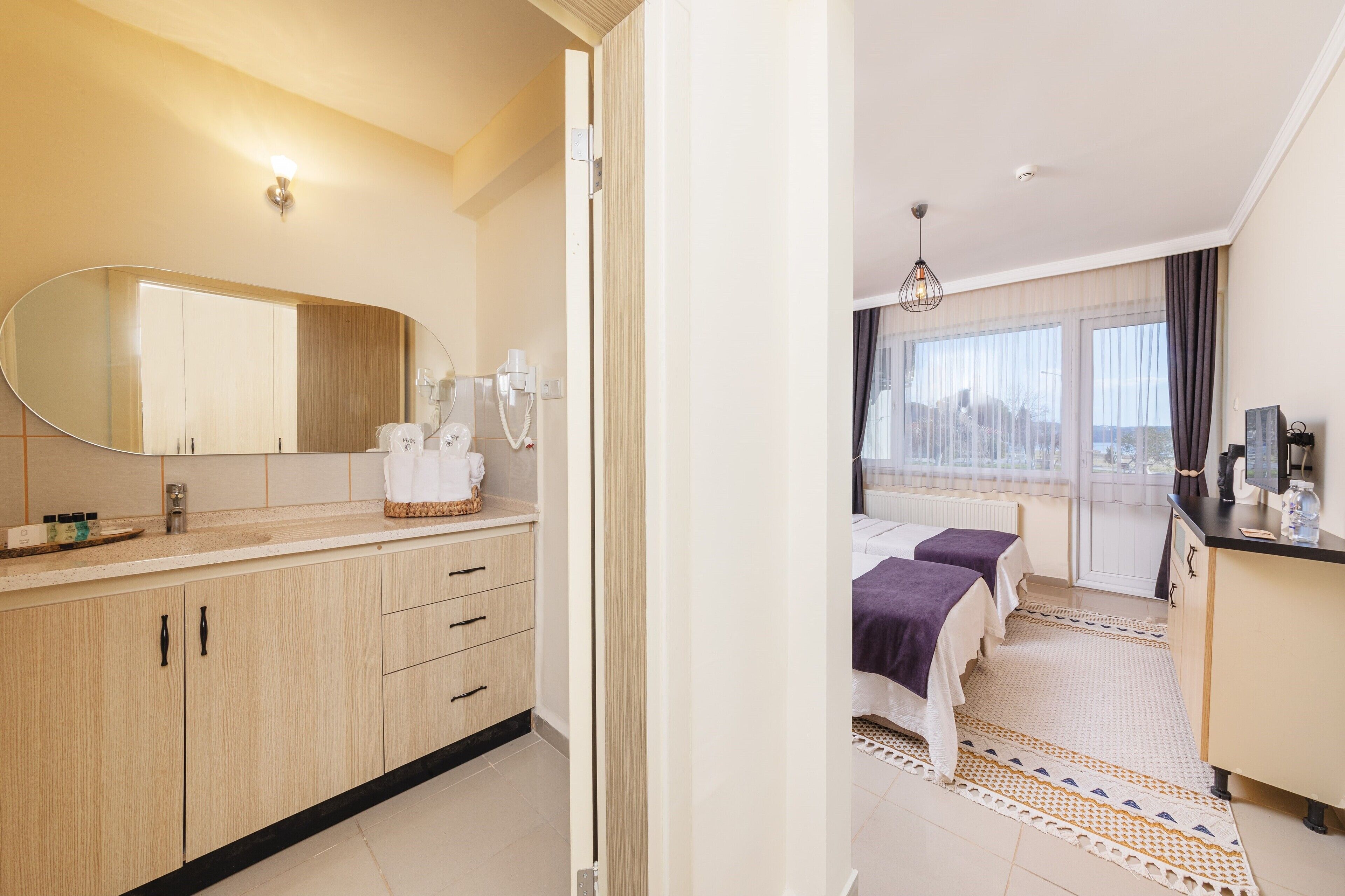 A modern hotel room with a large mirror, a vanity with toiletries, a bed with a view, and a small kitchenette.