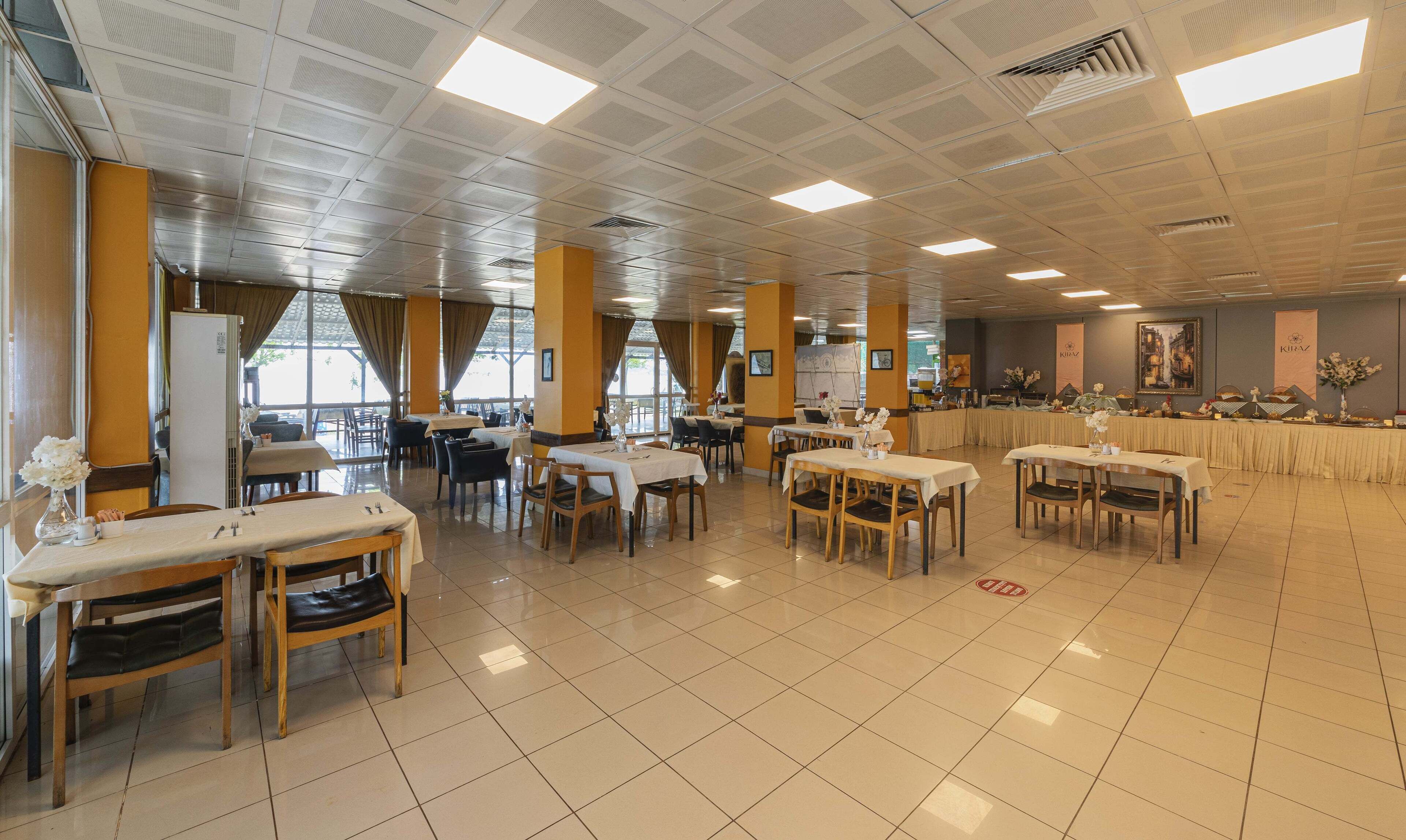 A spacious dining area with multiple tables and chairs, large windows, and a high ceiling with recessed lighting.