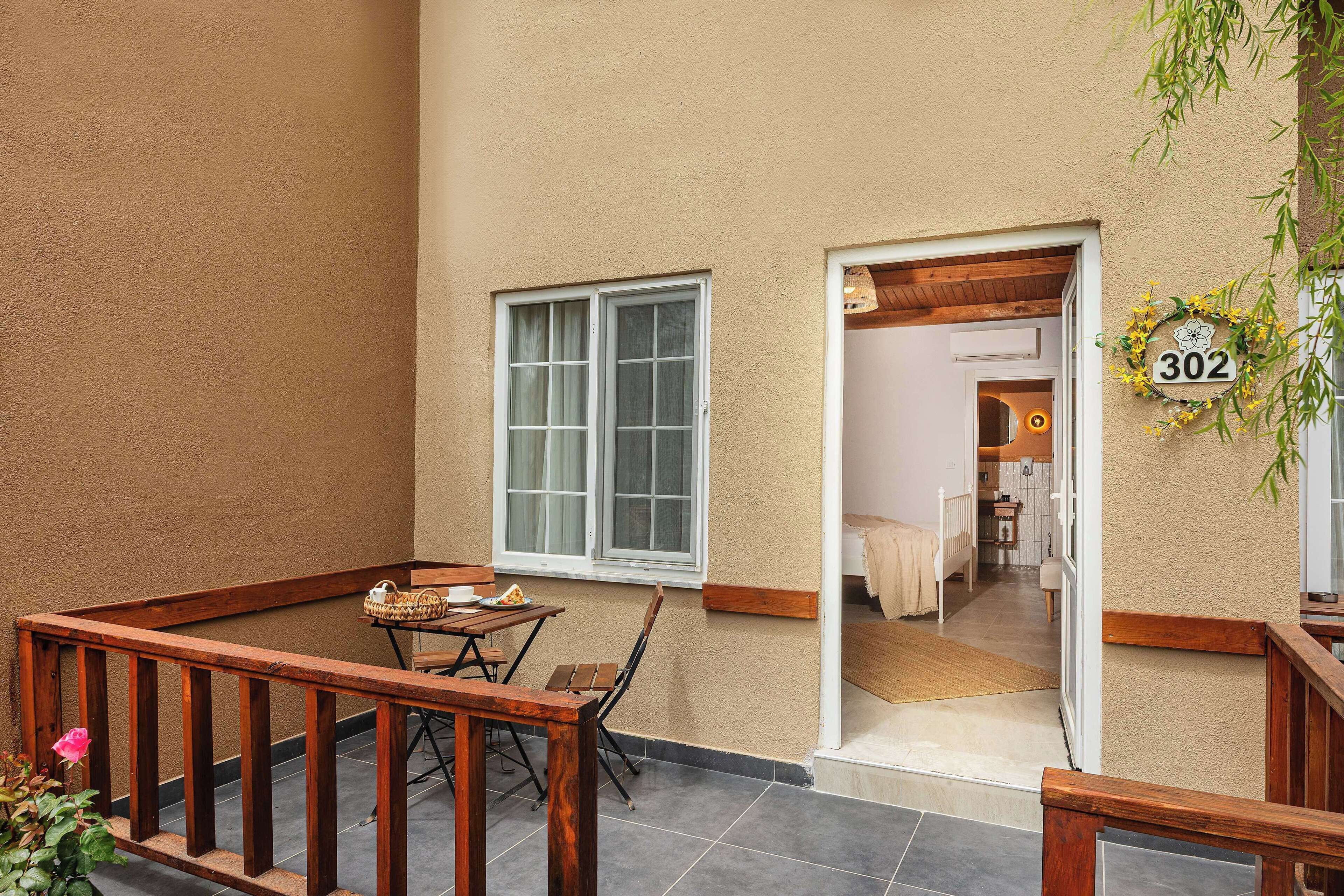 Une terrasse extérieure accueillante, avec une table et des chaises en bois, un petit jardin et une vue sur une chambre à coucher avec un lit et une table de chevet.