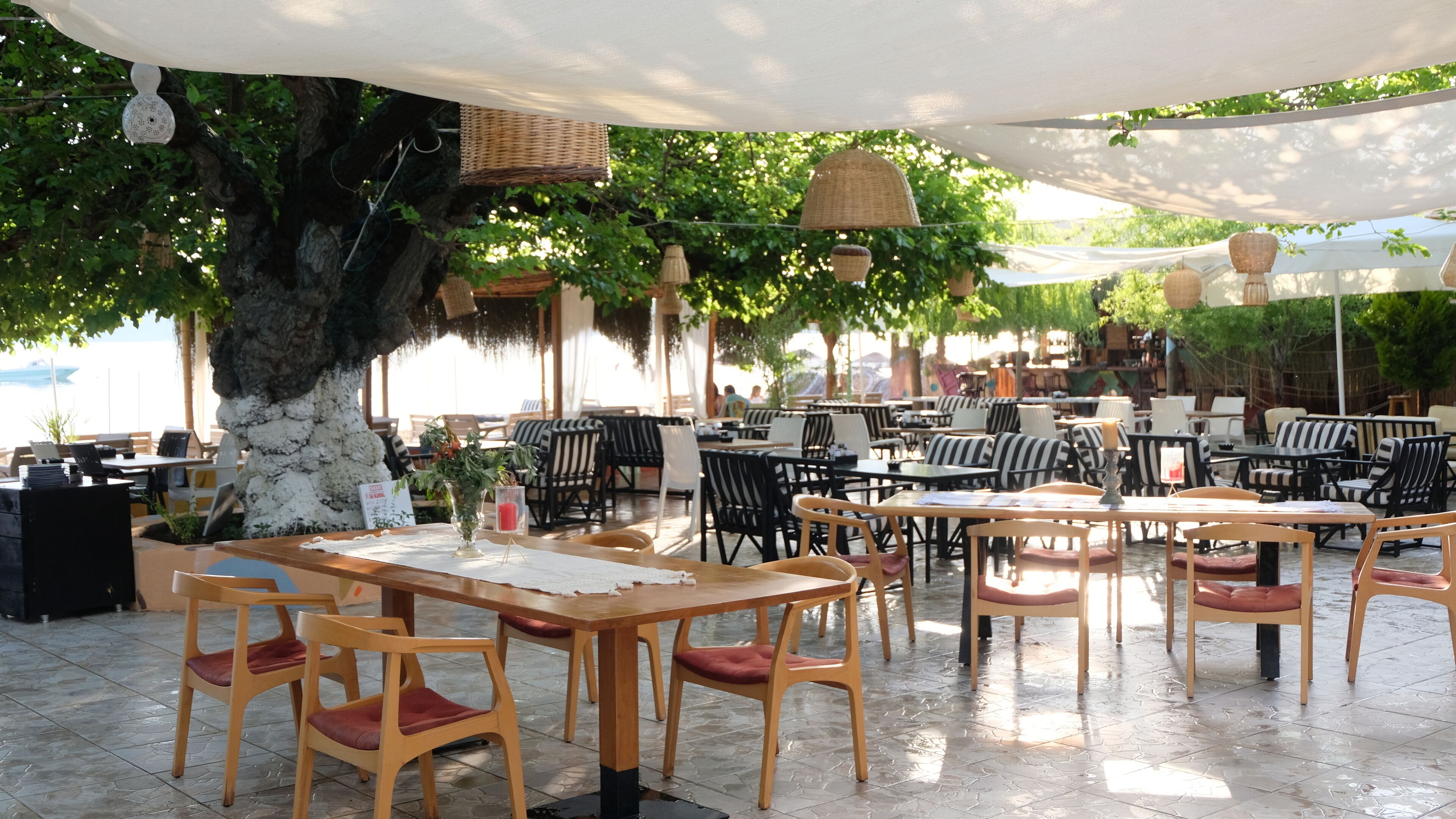 An outdoor cafe with wooden tables and chairs, surrounded by greenery and shaded by large umbrellas.