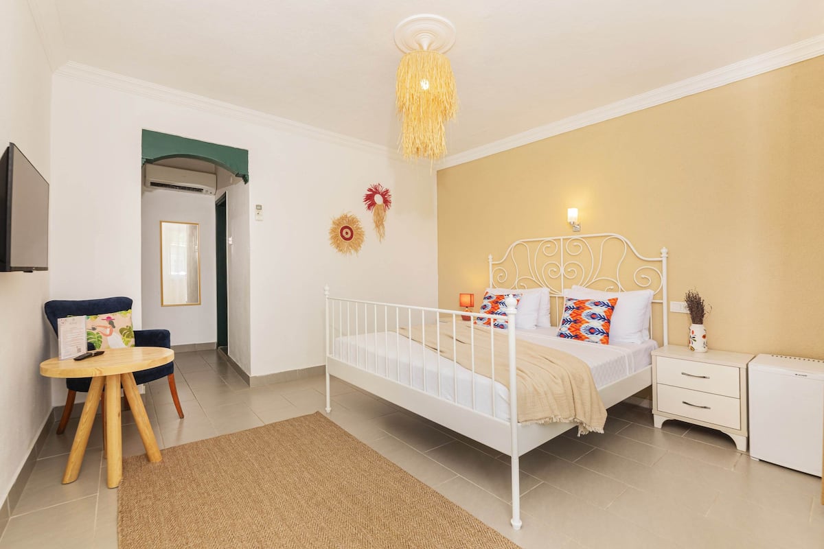 A bedroom with a white metal bed, bedside tables, a small white nightstand, a chair with a table, and a decorative ceiling light.