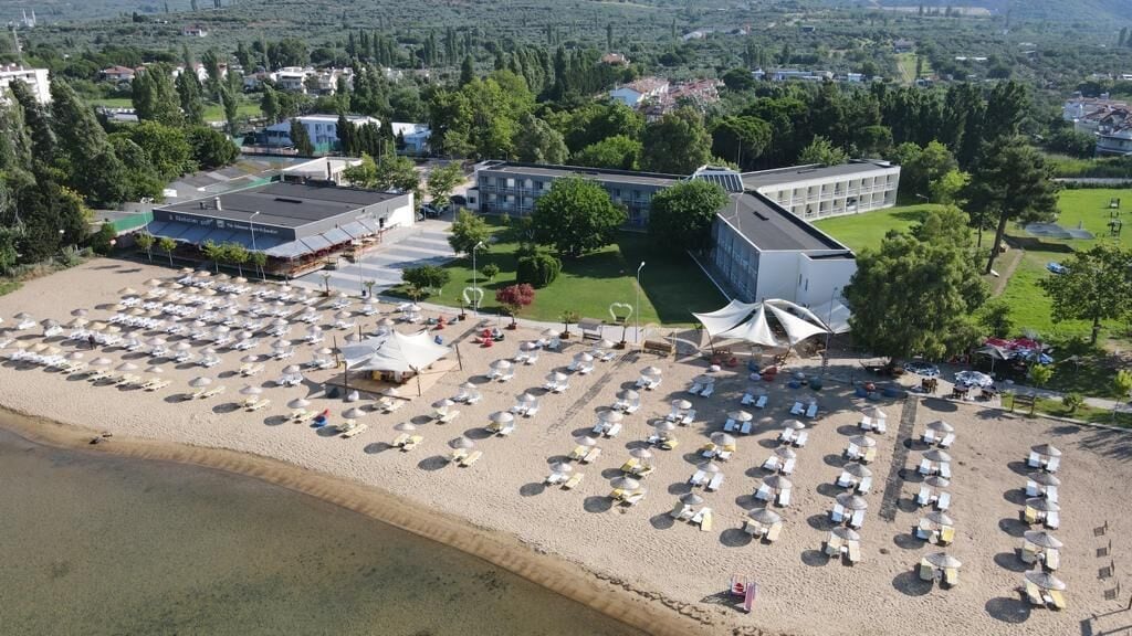 Eine Vogelperspektive zeigt einen Strand mit vielen Liegestühlen und Sonnenschirmen, ein großes Gebäude mit flachem Dach und einen Parkplatz.