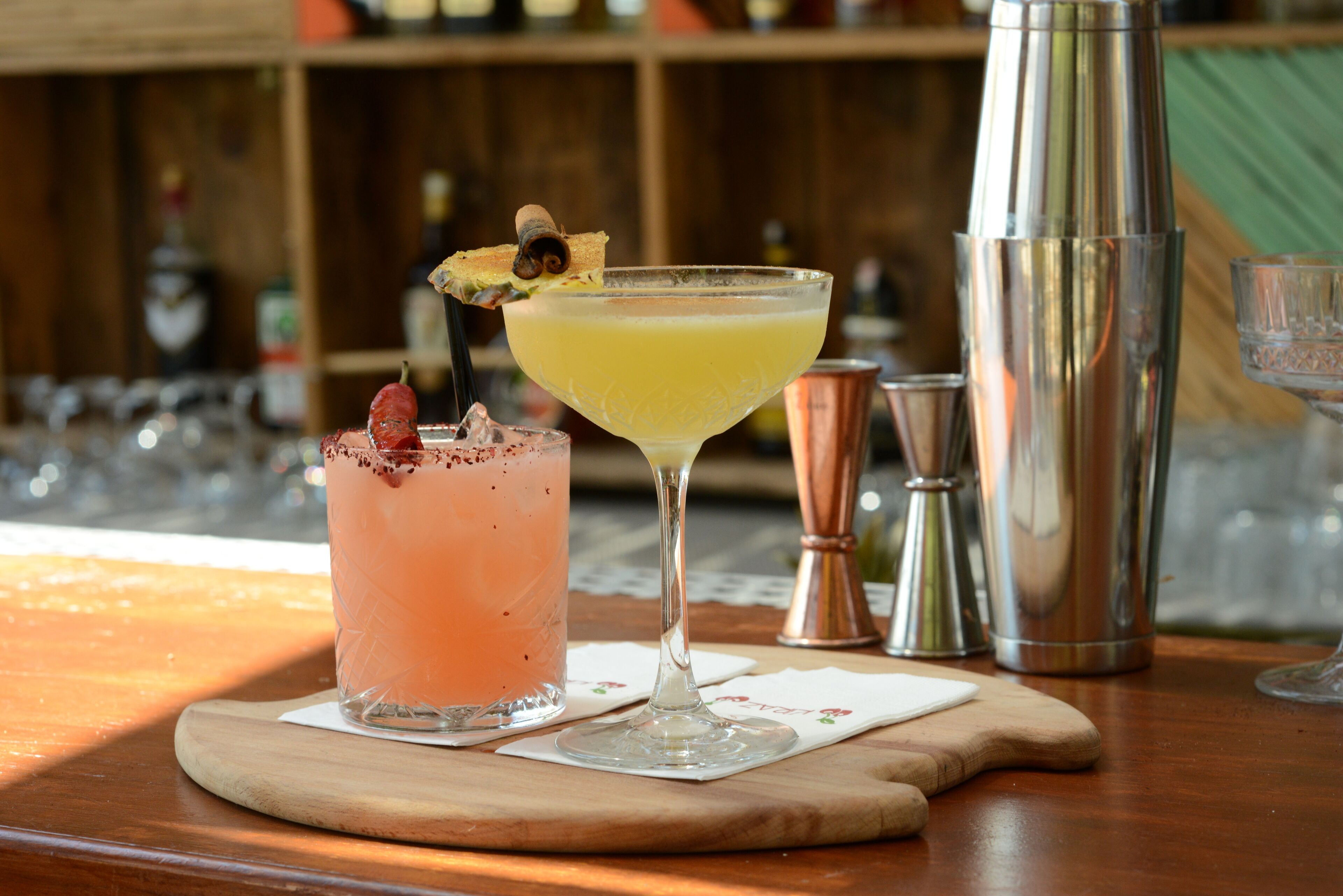 Two cocktails on a wooden bar counter with a shaker and copper jigger in the background.