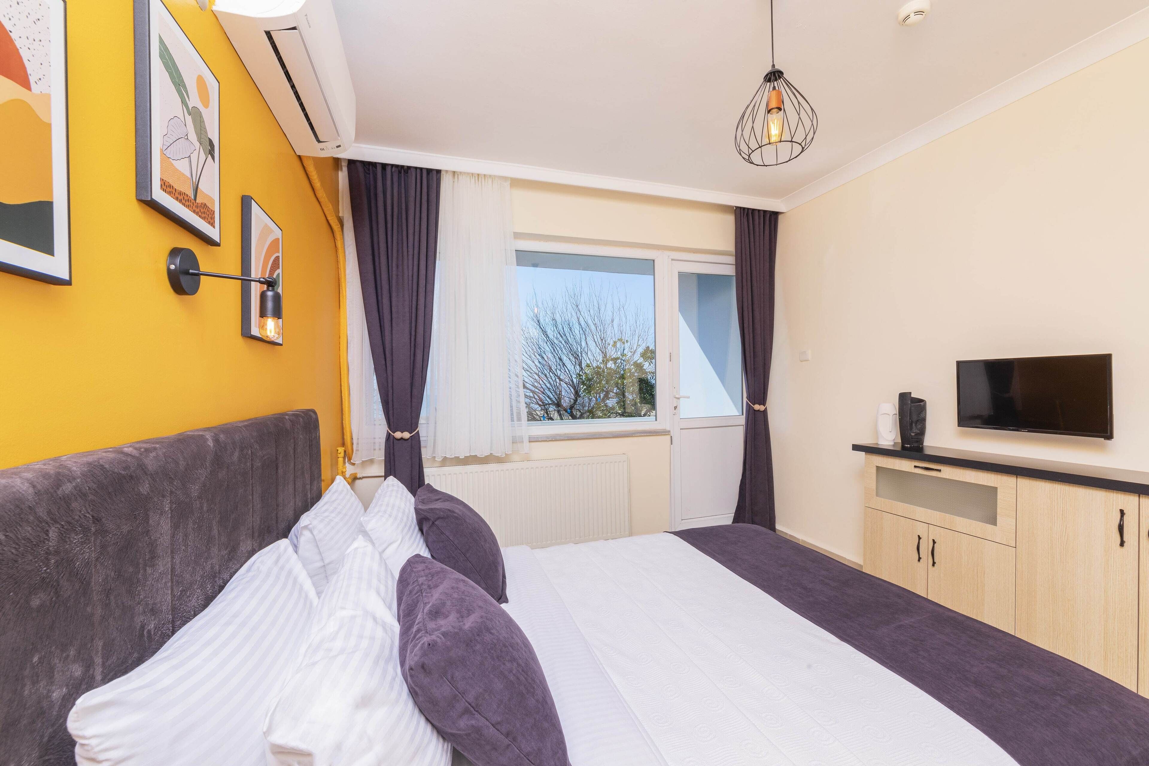 A hotel room with a bed, a TV, a window with curtains, and artwork on the wall.