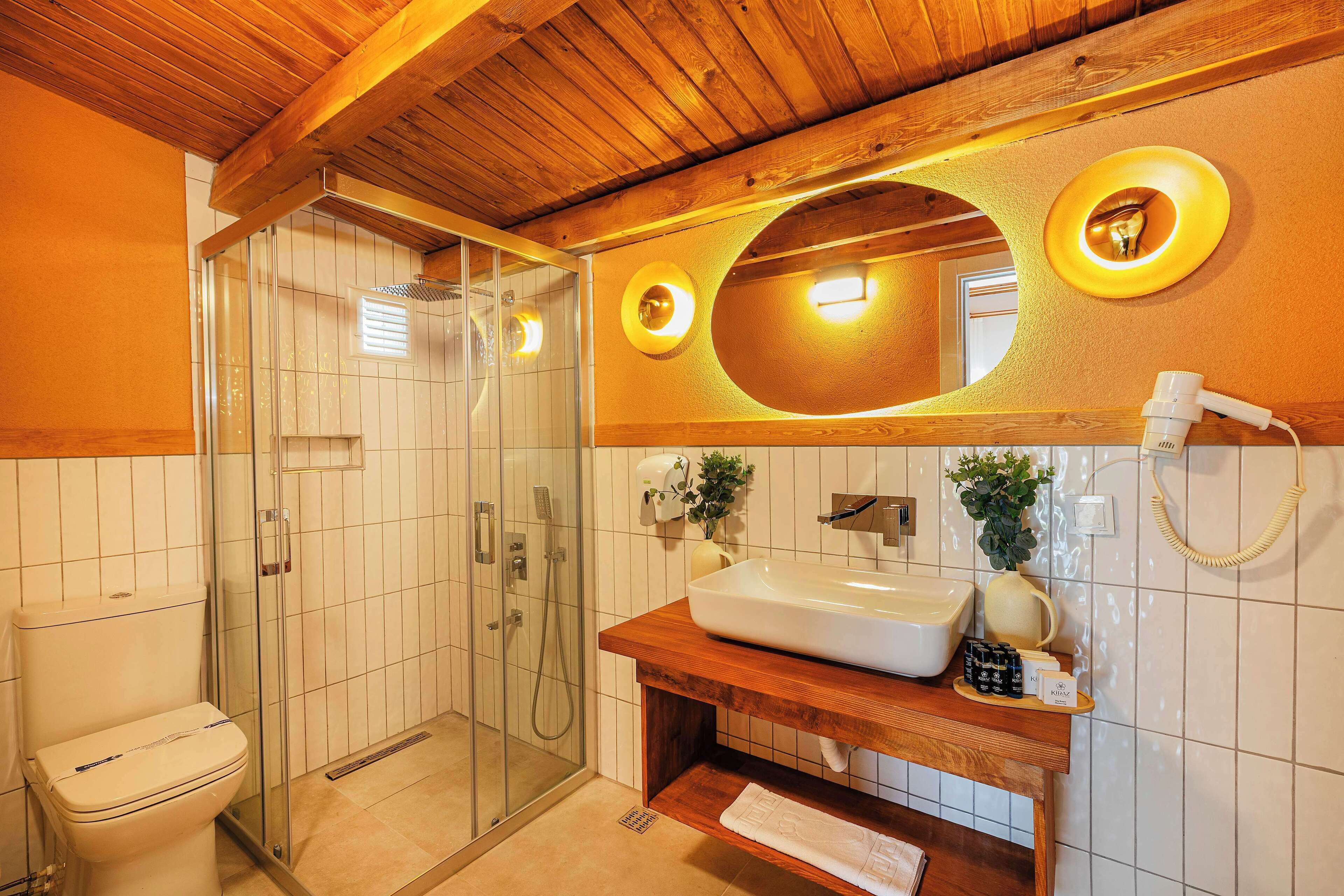 Een badkamer met een houten plafond, een douchecabine van glas, een witte wastafel en een spiegel met een ronde lamp.