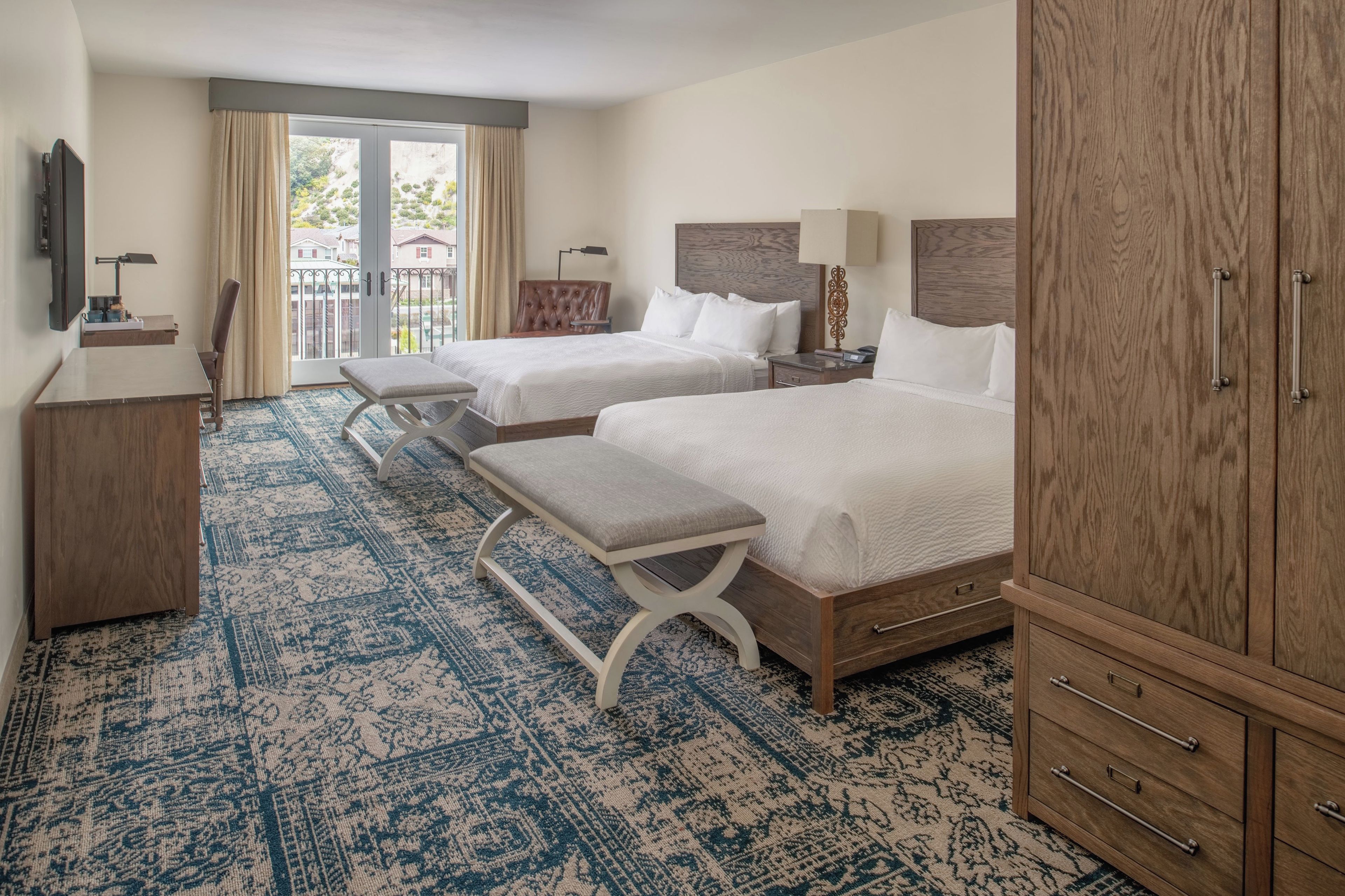 Habitación de hotel con dos camas, un escritorio, una silla, un armario y vistas al exterior.