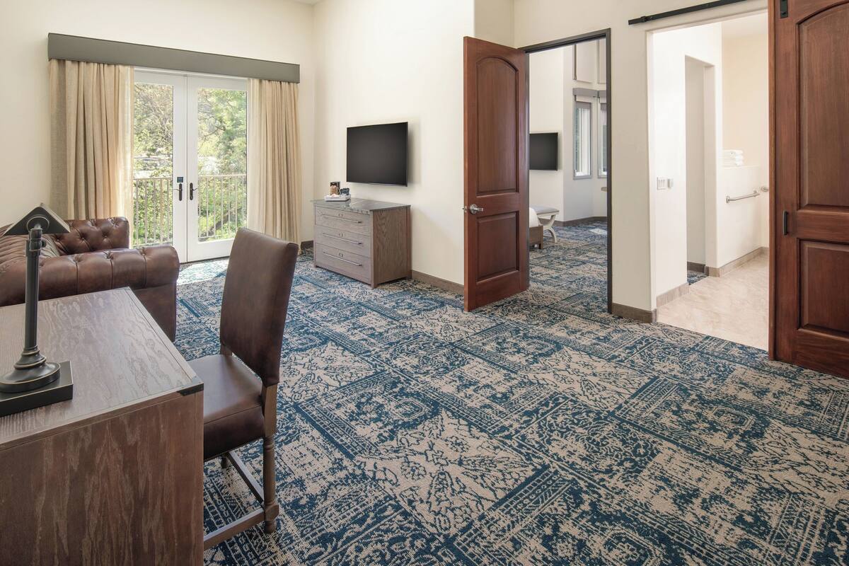 Une chambre d’hôtel dotée d’une grande fenêtre, d’un téléviseur à écran plat, d’un bureau en bois, d’un fauteuil en cuir marron et d’un tapis à motifs.