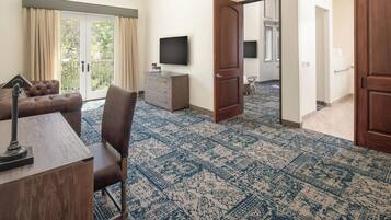 Habitación de hotel con un ventanal grande, un televisor de pantalla plana, un escritorio de madera, una silla de cuero marrón y una alfombra estampada.