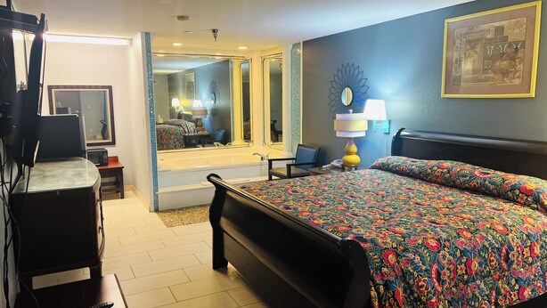 A hotel room with a large bed, a desk with a lamp, a chair, a television, and a bathroom visible through an open door. - The Shades Motel (Baton Rouge)