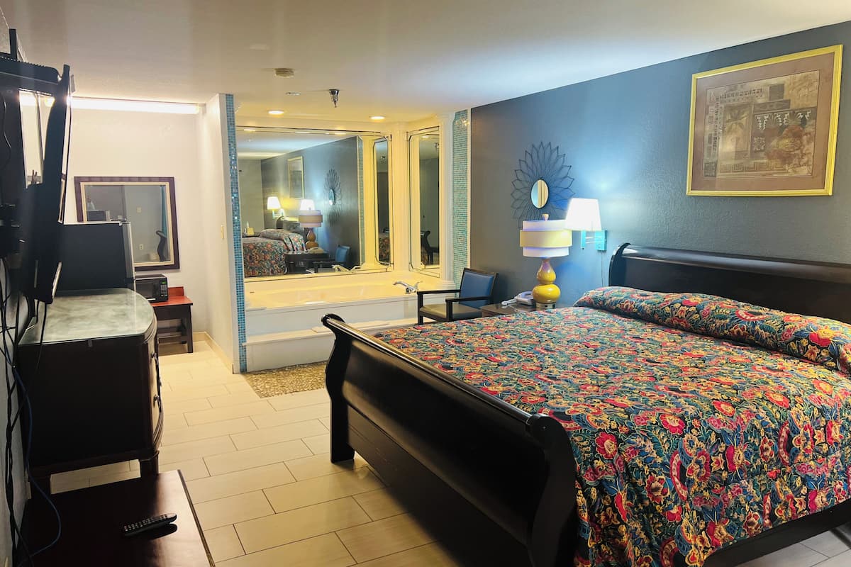 A hotel room with a large bed, a desk with a lamp, a chair, a television, and a bathroom visible through an open door.