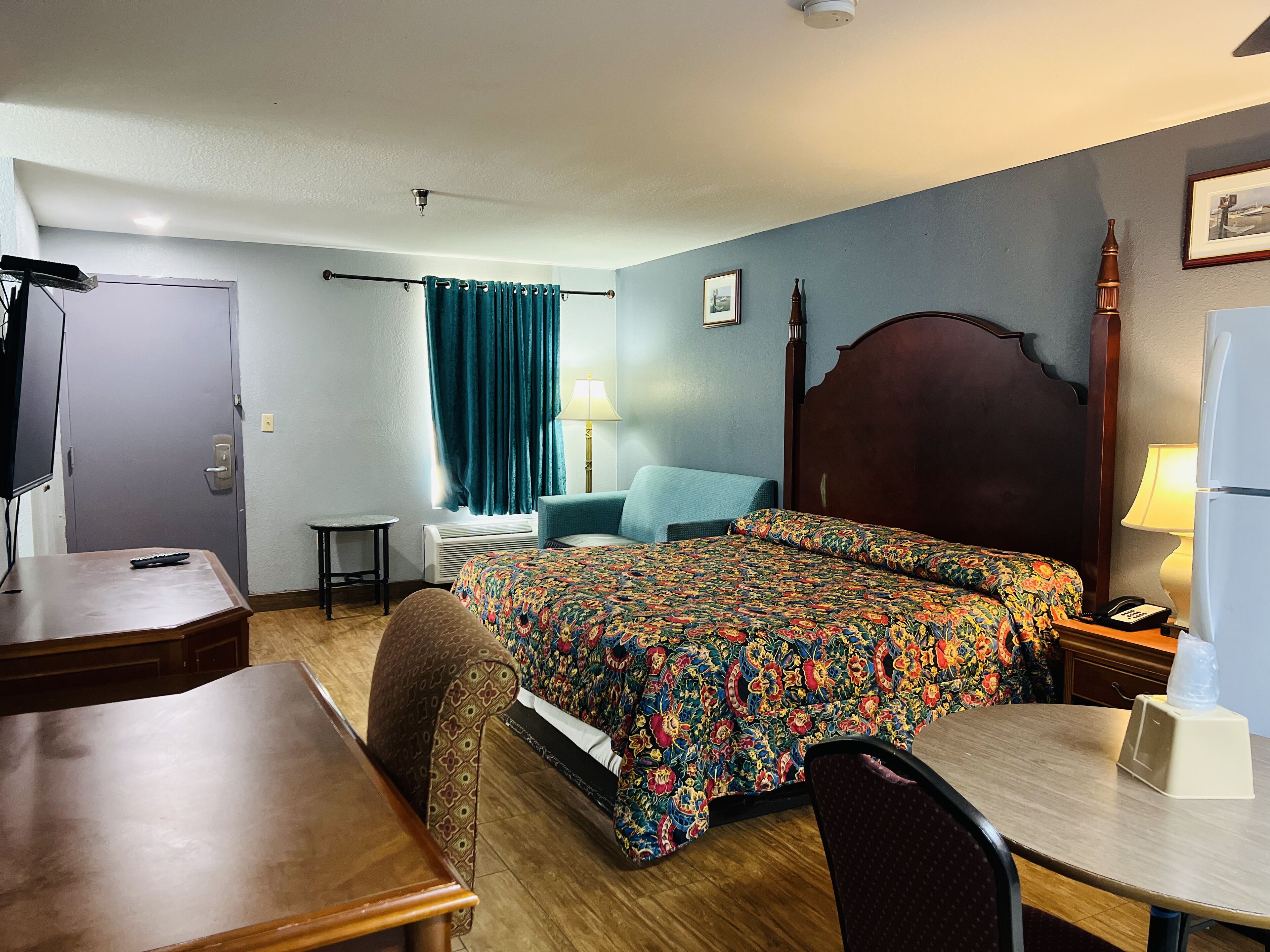 A hotel room with a bed, a desk, a chair, a table, a lamp, a refrigerator, and a window with curtains.