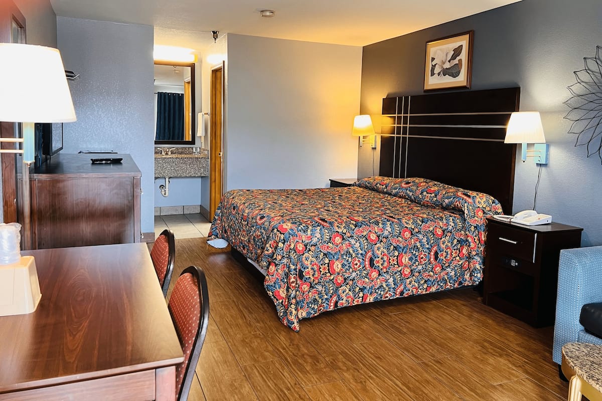 A hotel room with a bed, a desk, a chair, a nightstand, a lamp, a mirror, and a bathroom visible through an open door.