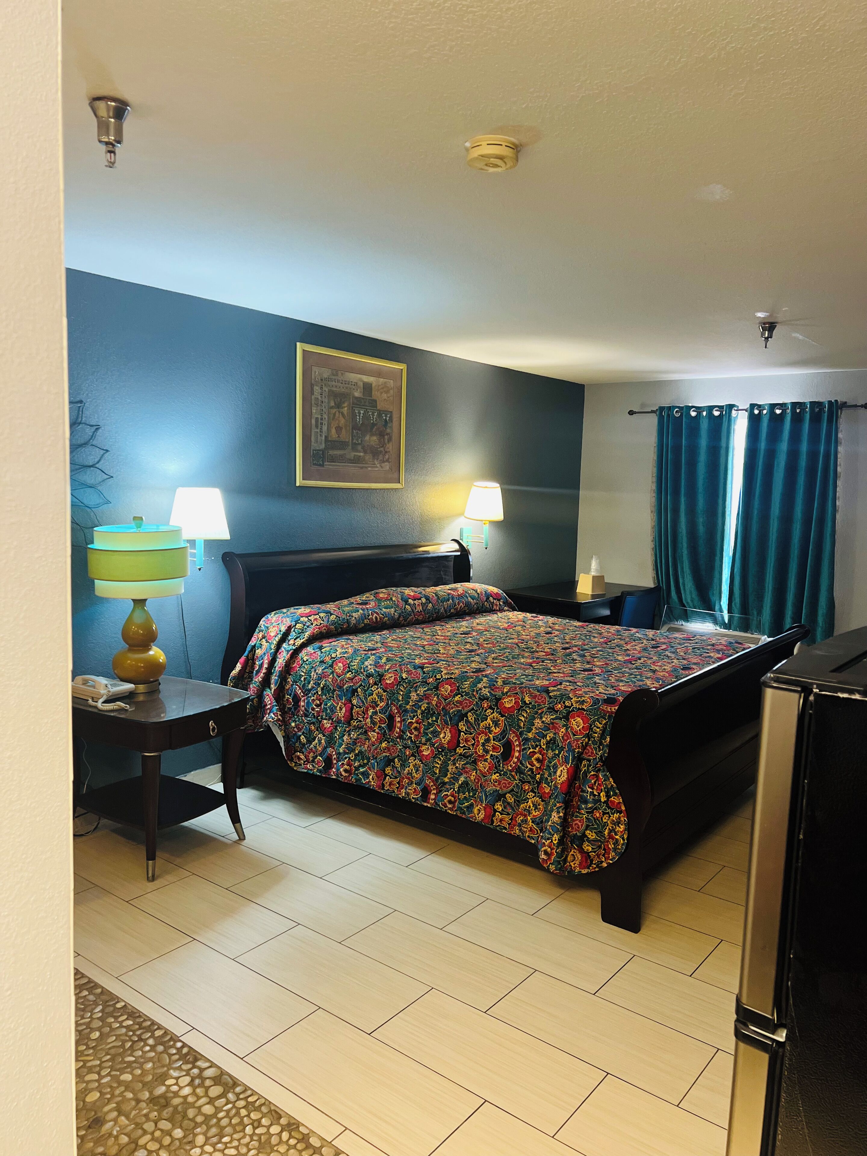 Ein Hotelzimmer mit einem Bett, zwei Nachttischen, einem Schreibtisch mit Lampe, einem Wandbild und einem Deckenventilator.