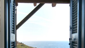 Vista desde una ventana con contraventanas abiertas, con paisaje costero y una mesa con un bol de fruta y una botella.