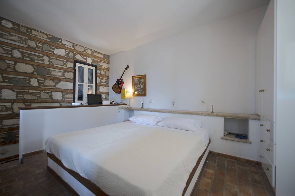 A bedroom with a stone wall, a bed, a guitar, and a small shelf with a lamp.