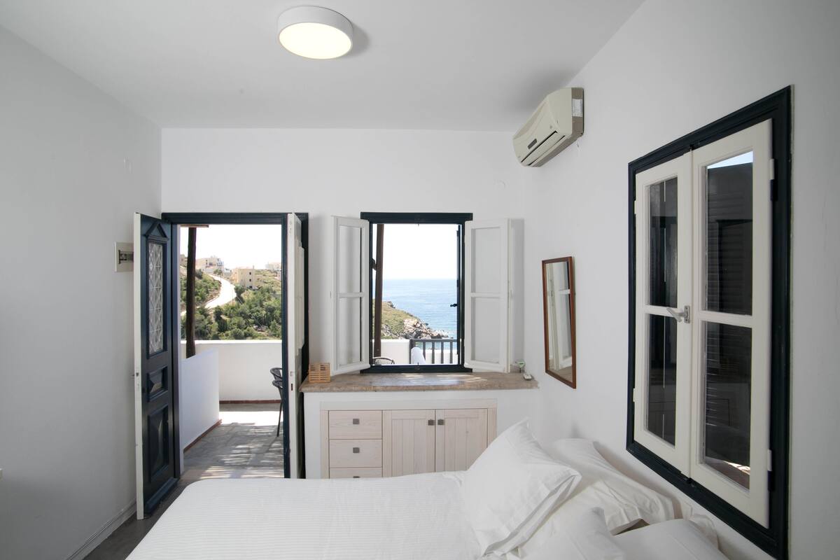 A bedroom with a bed, a window with a view of the sea, a balcony, and an air conditioning unit.