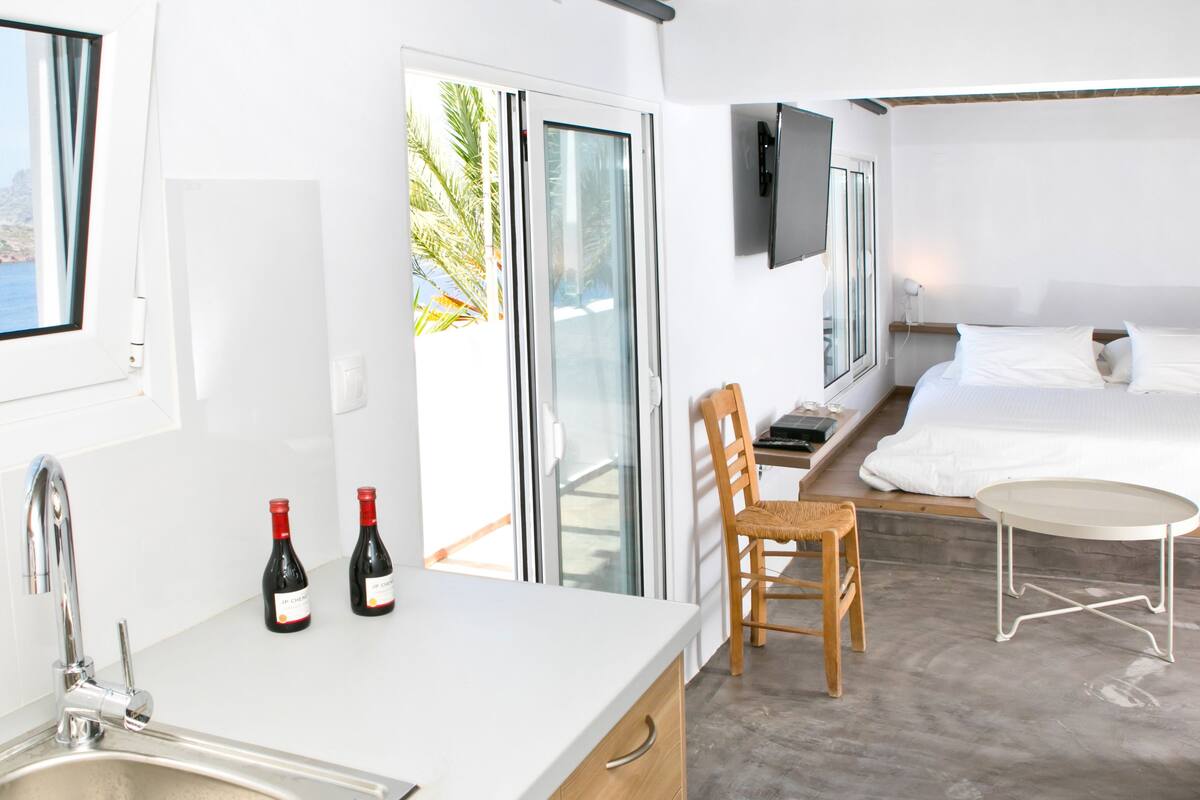 A modern hotel room with a large window, a bed, a small table, a chair, and a sink.