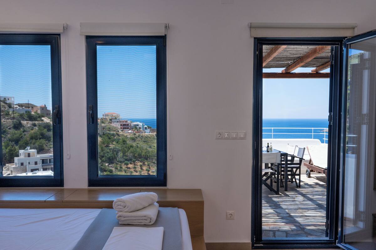 A bedroom with a large bed, two windows, and a balcony with a view of the sea.