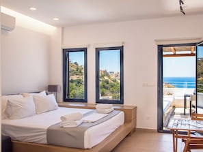 A bedroom with a bed, a bedside table, a lamp, and a view of a coastal landscape through large windows. - Blue Dawn Kalymnos (Kalymnos)