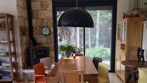 A cozy dining area with a wooden table, chairs, and a hanging light fixture. A fireplace and a bookshelf are visible in the background.