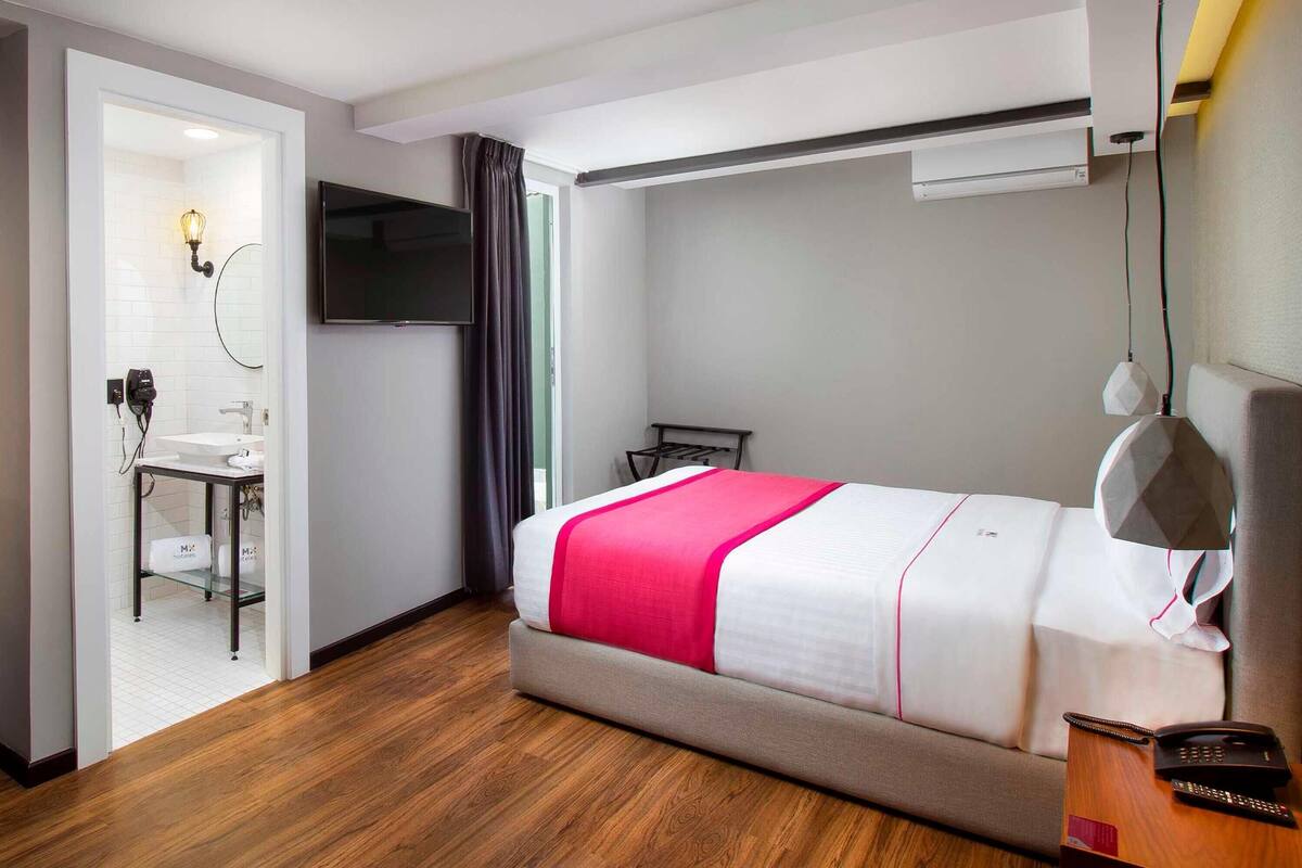 A hotel room with a bed, a desk, a chair, a TV, and a bathroom visible through an open door.