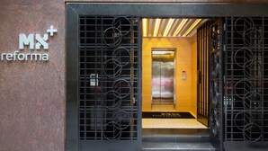 Entrada de hotel con diseño moderno, que presenta una reja metálica con motivos circulares, un ascensor a la vista y un cartel que dice "MX reforma".
