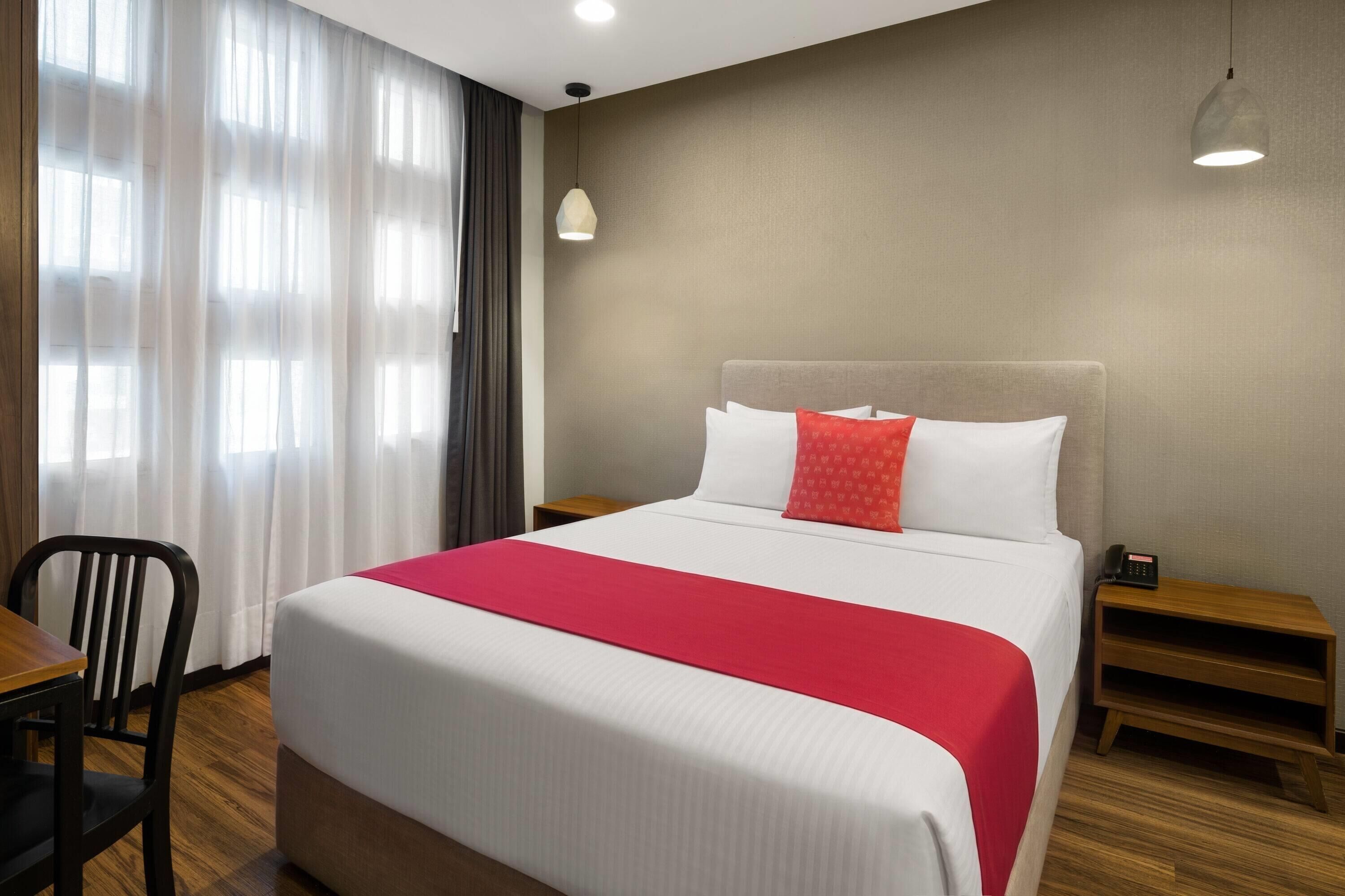 Quarto de hotel com cama, cadeira, mesa de cabeceira, luminária e janela com cortinas.