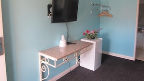 Una habitación con una televisión de pantalla plana montada en la pared, un pequeño escritorio blanco con un control remoto, un dispensador de gel antibacterial y una planta en maceta con flores.