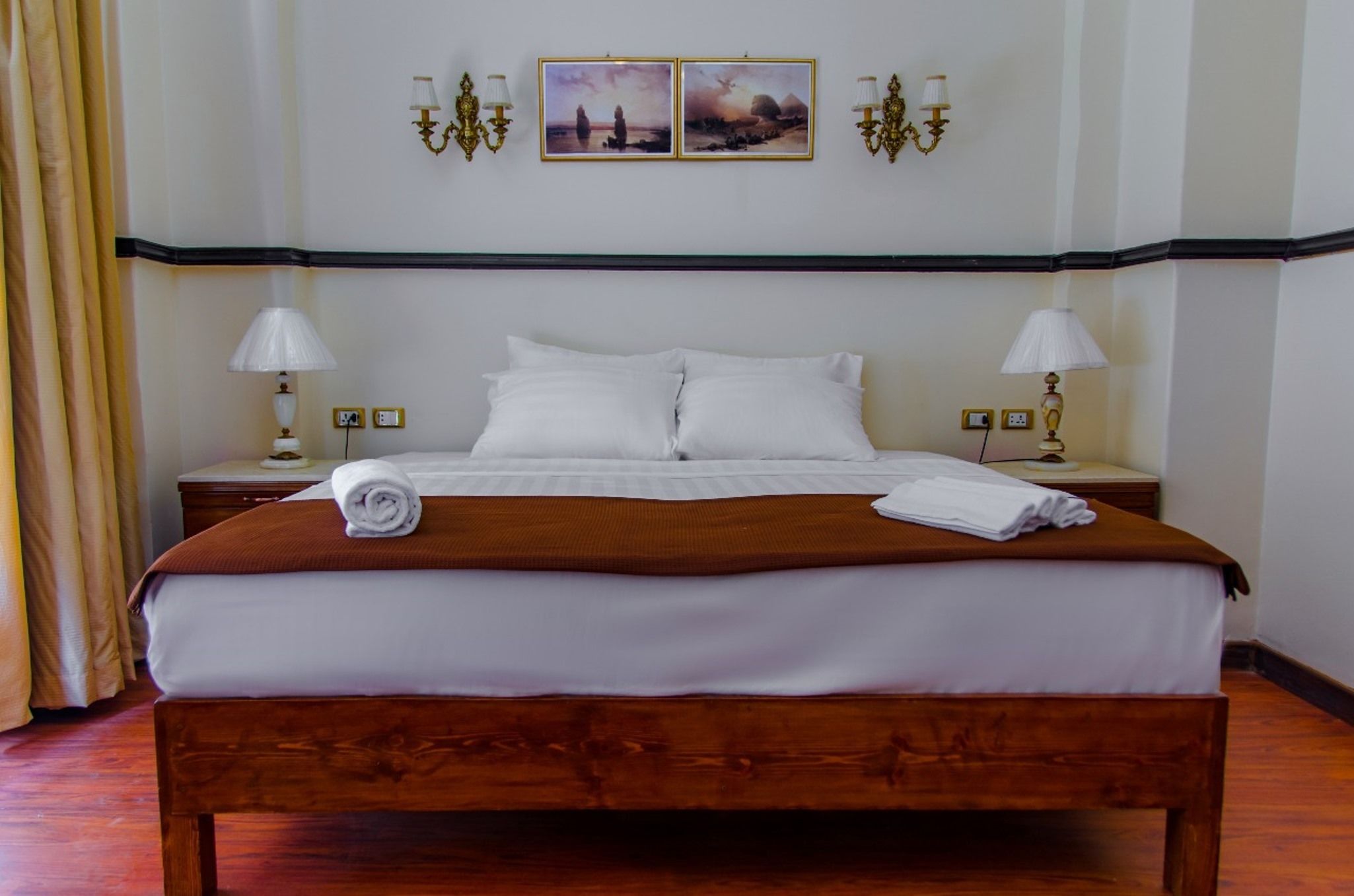 A neatly made bed with white linens and a brown blanket, two bedside lamps, and two framed pictures on the wall.