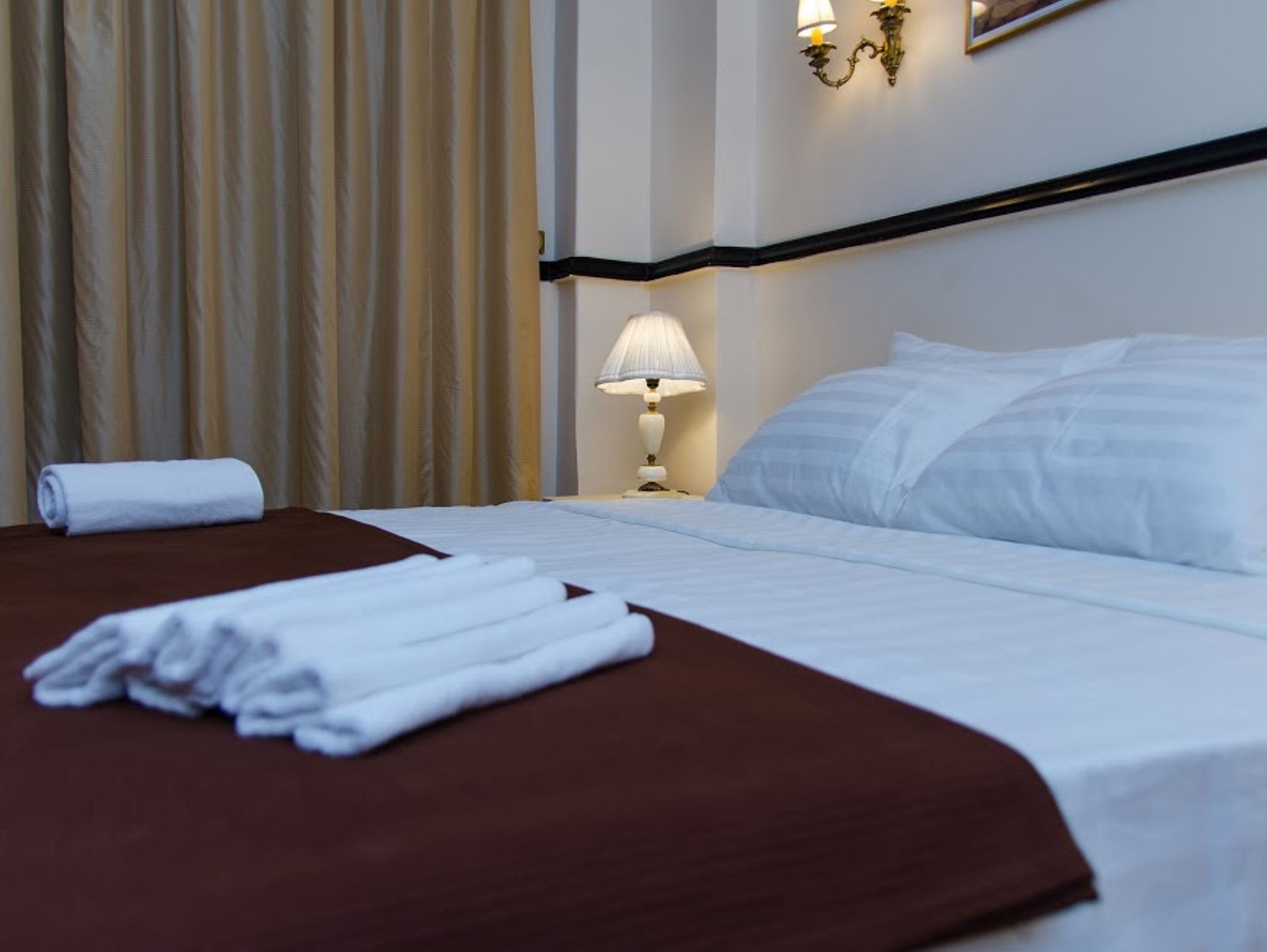 A neatly made bed with white linens, a brown bedspread, and a rolled white towel on the side.