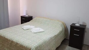 A neatly made bed with a green patterned bedspread and folded white towels. There are two bedside tables, one with a lamp and the other with a small white object on top. - La Casa (San Lorenzo)