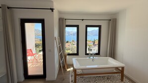 A bathroom with a bathtub, two windows with a view of a lake and mountains, and a door leading to an outdoor area with a red chair. - Gala Selimiye (Marmaris)