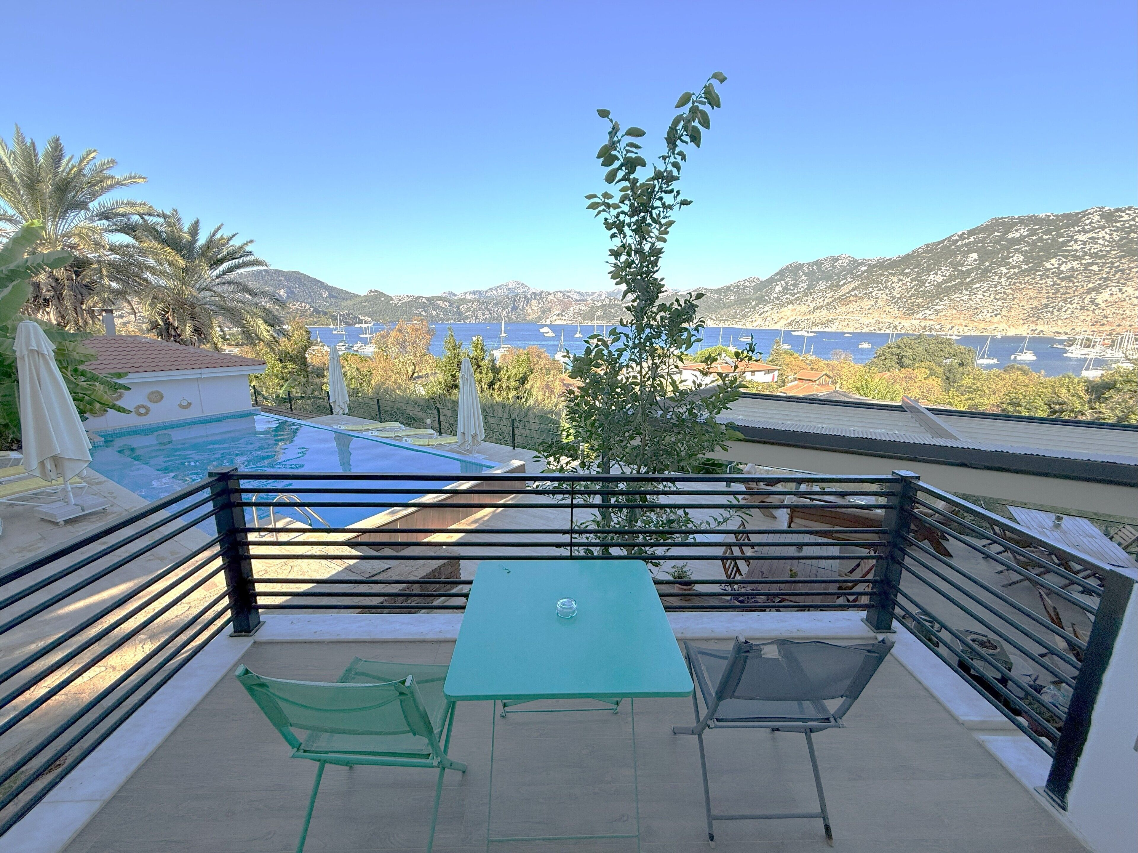 A balcony with a pool, two chairs, a table, and a view of a mountainous landscape and the sea.