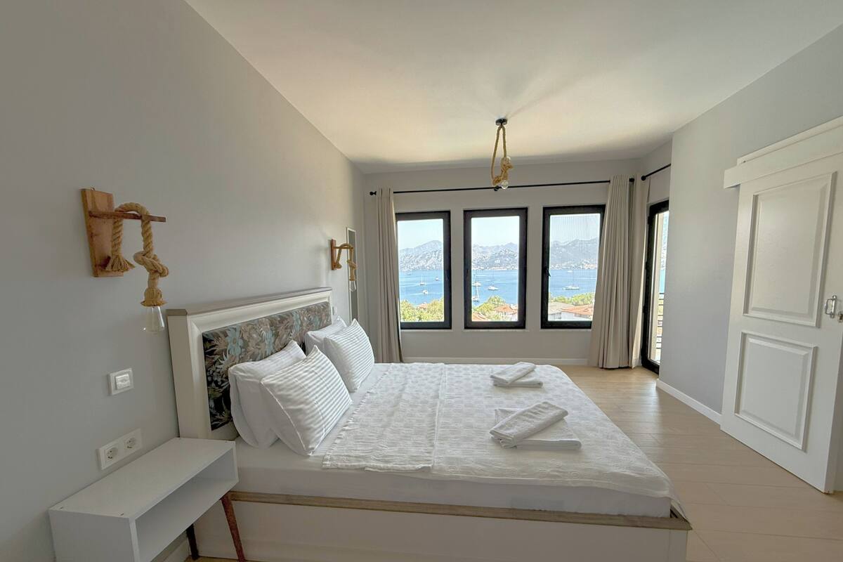 A modern bedroom with a large bed, a bedside table, a view of the sea through large windows, and a ceiling light.