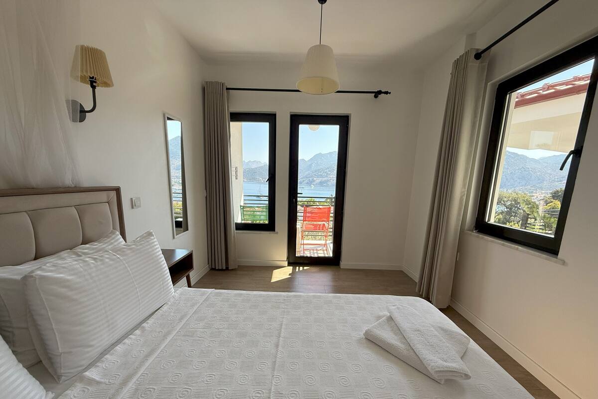 A bedroom with a large bed, a nightstand, a lamp, a window with a view of the sea, and a balcony with chairs.