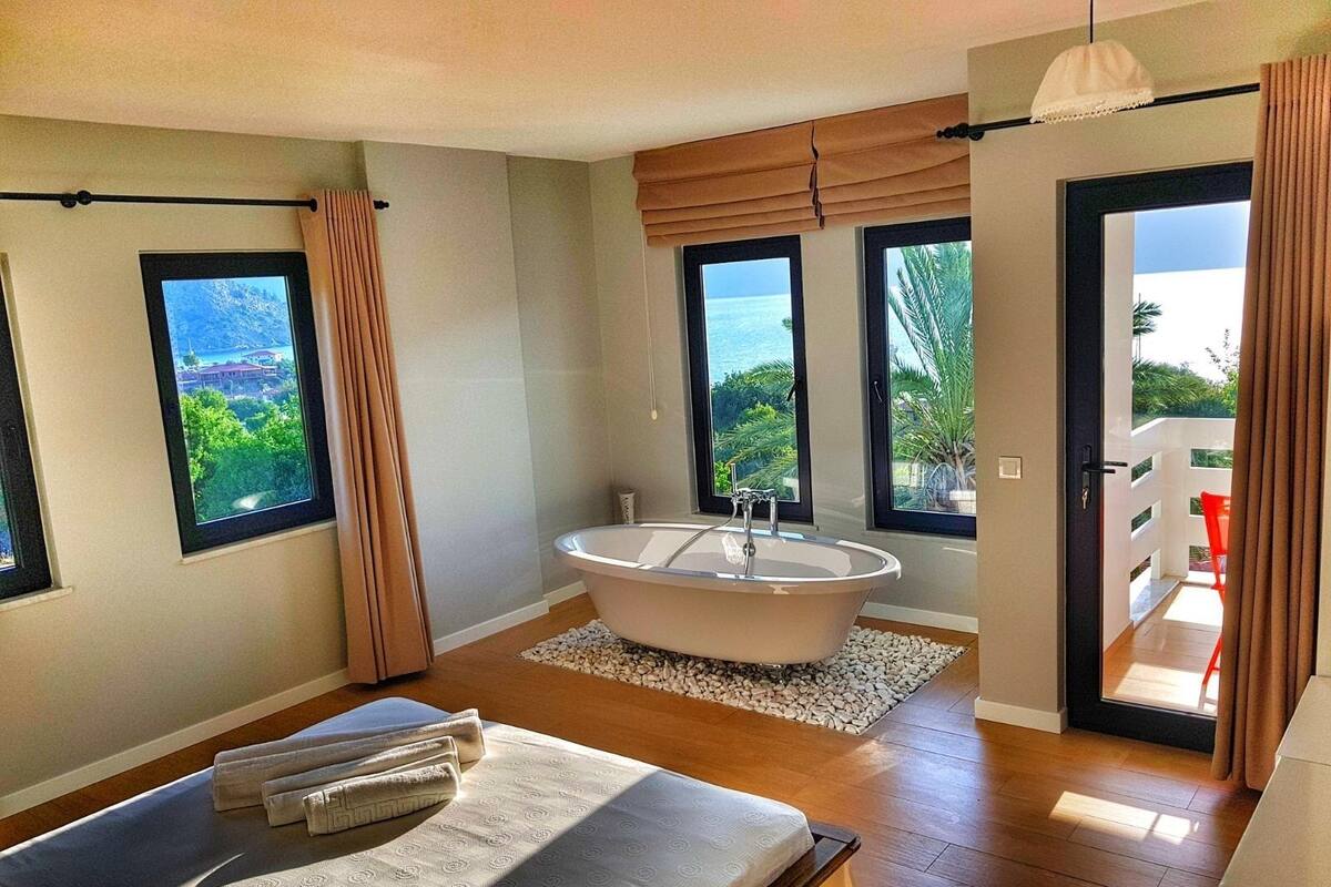 A modern bedroom with a large bed, a freestanding bathtub, and a balcony with a view of the ocean.