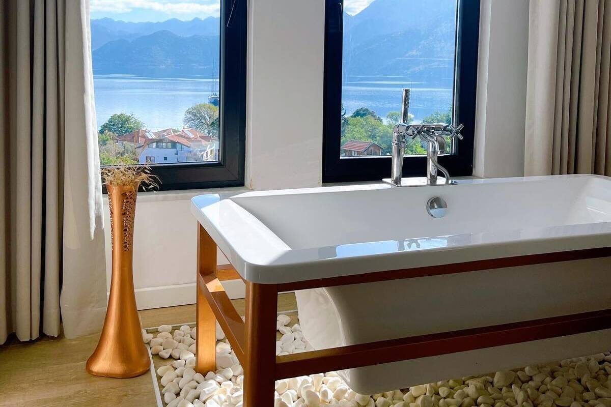 A modern bathroom with a large bathtub, two windows with a scenic view, and a decorative vase.