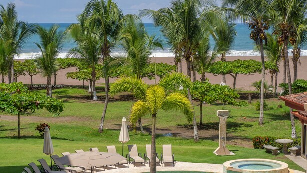 A pool area with lounge chairs and umbrellas, surrounded by palm trees and a view of the beach and ocean. - Tres Regalos N302 (Jacó)