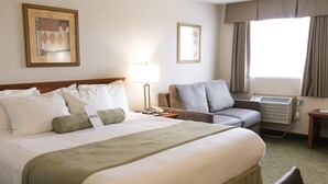 Hotellihuone, jossa on suuri sänky, harmaa nojatuoli, kirjoituspöytä puhelimineen ja ikkuna, jossa on verhot.