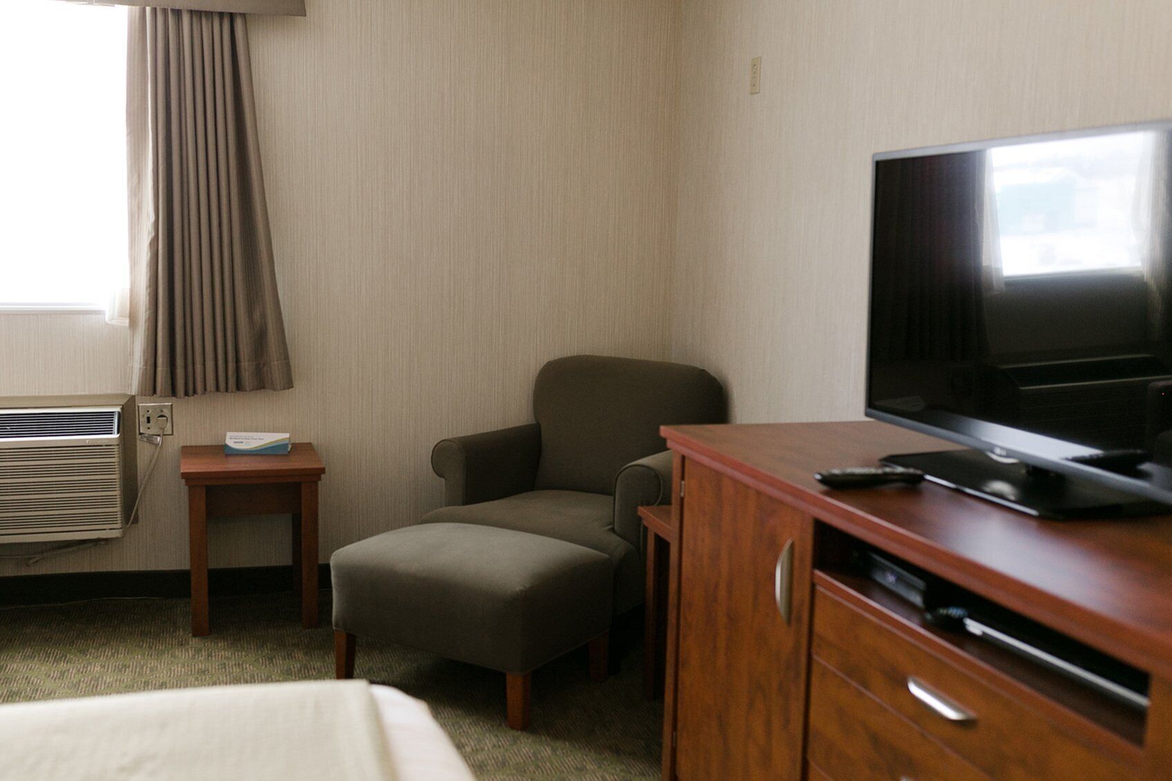 Quarto de hotel com uma cama, televisão, escrivaninha de madeira, cadeira e uma pequena mesa com controle remoto.