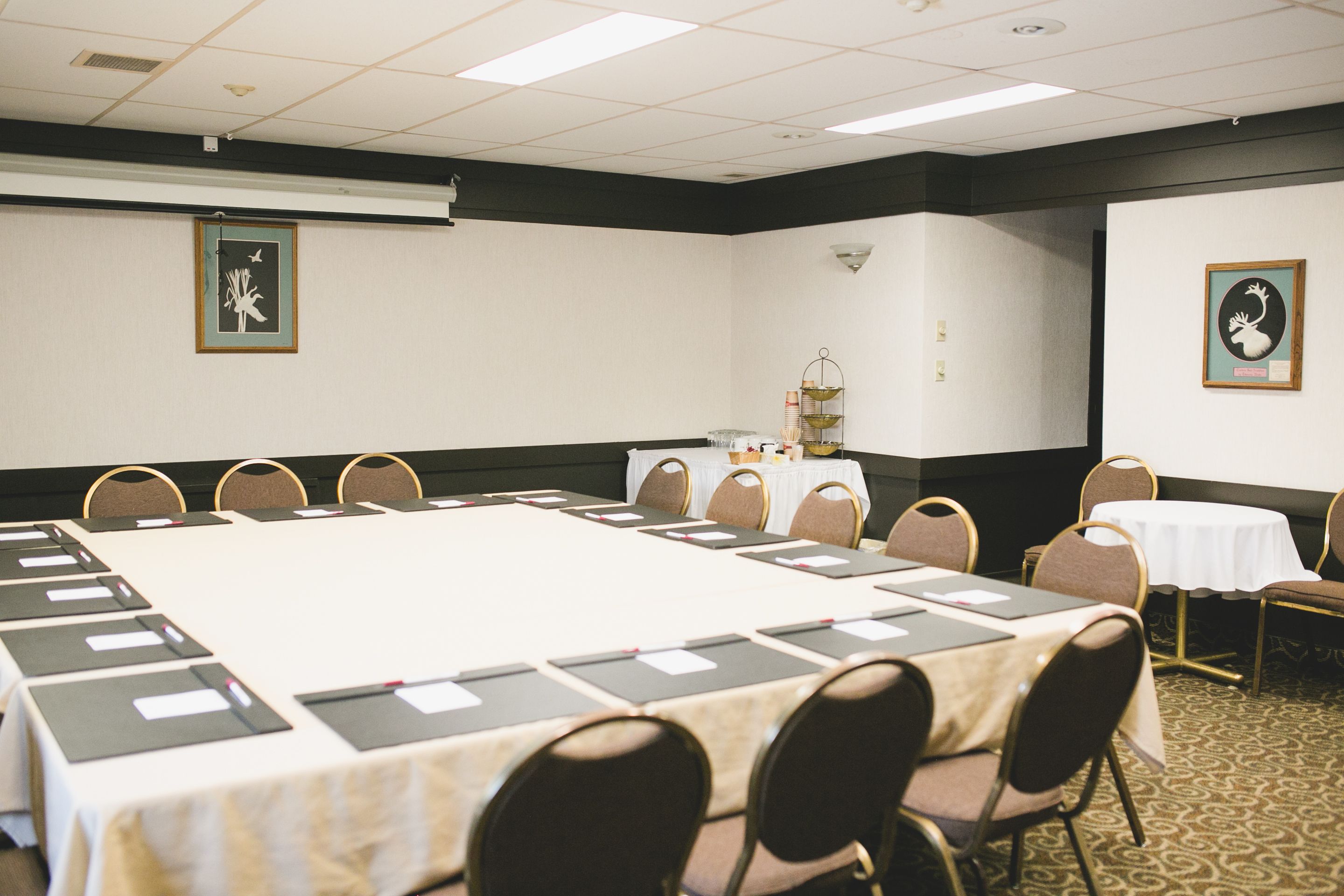 A conference room with a large table, chairs, a projector screen, and framed artwork on the walls.