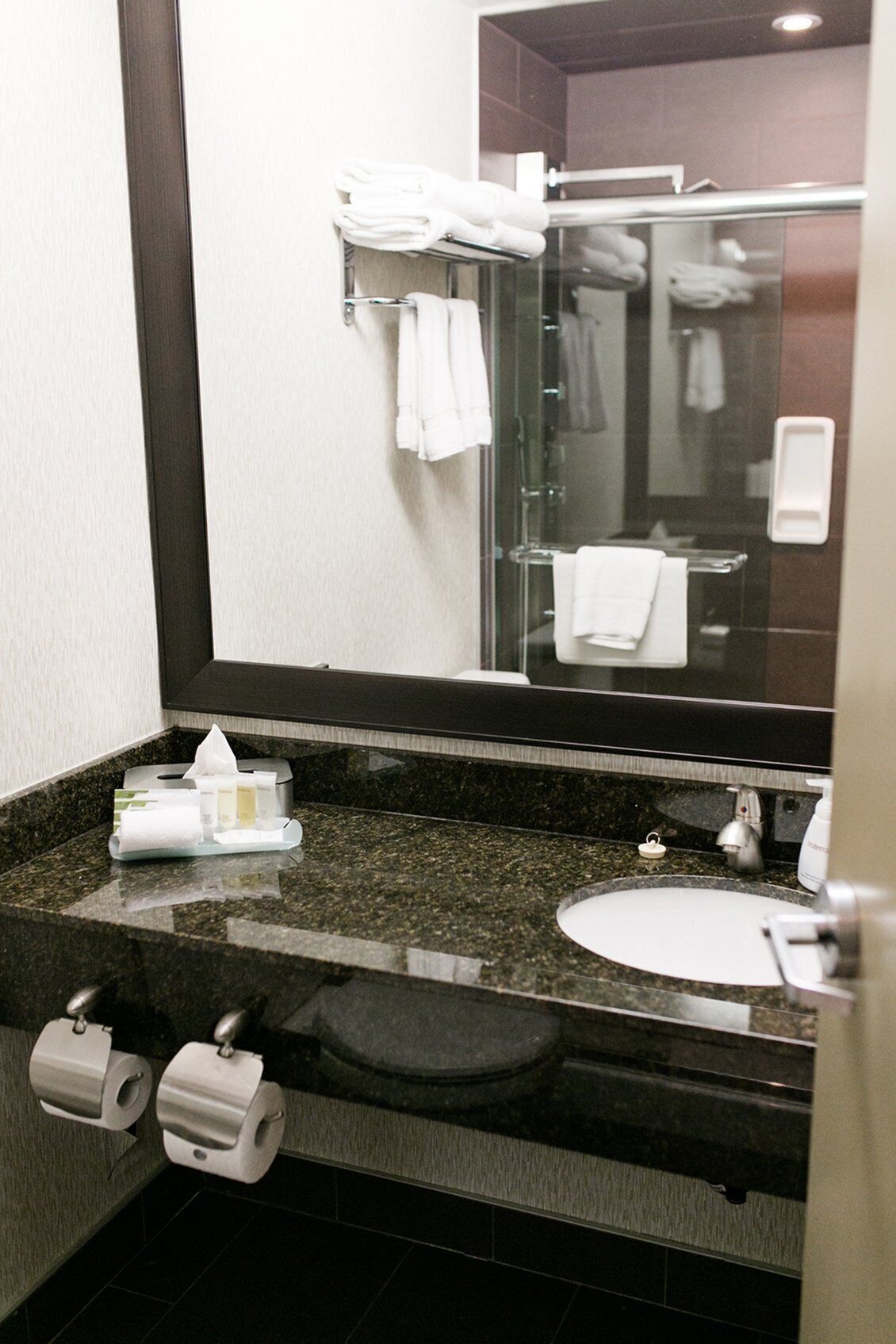 A hotel bathroom with a granite countertop, a sink, a mirror, and a glass shower enclosure.