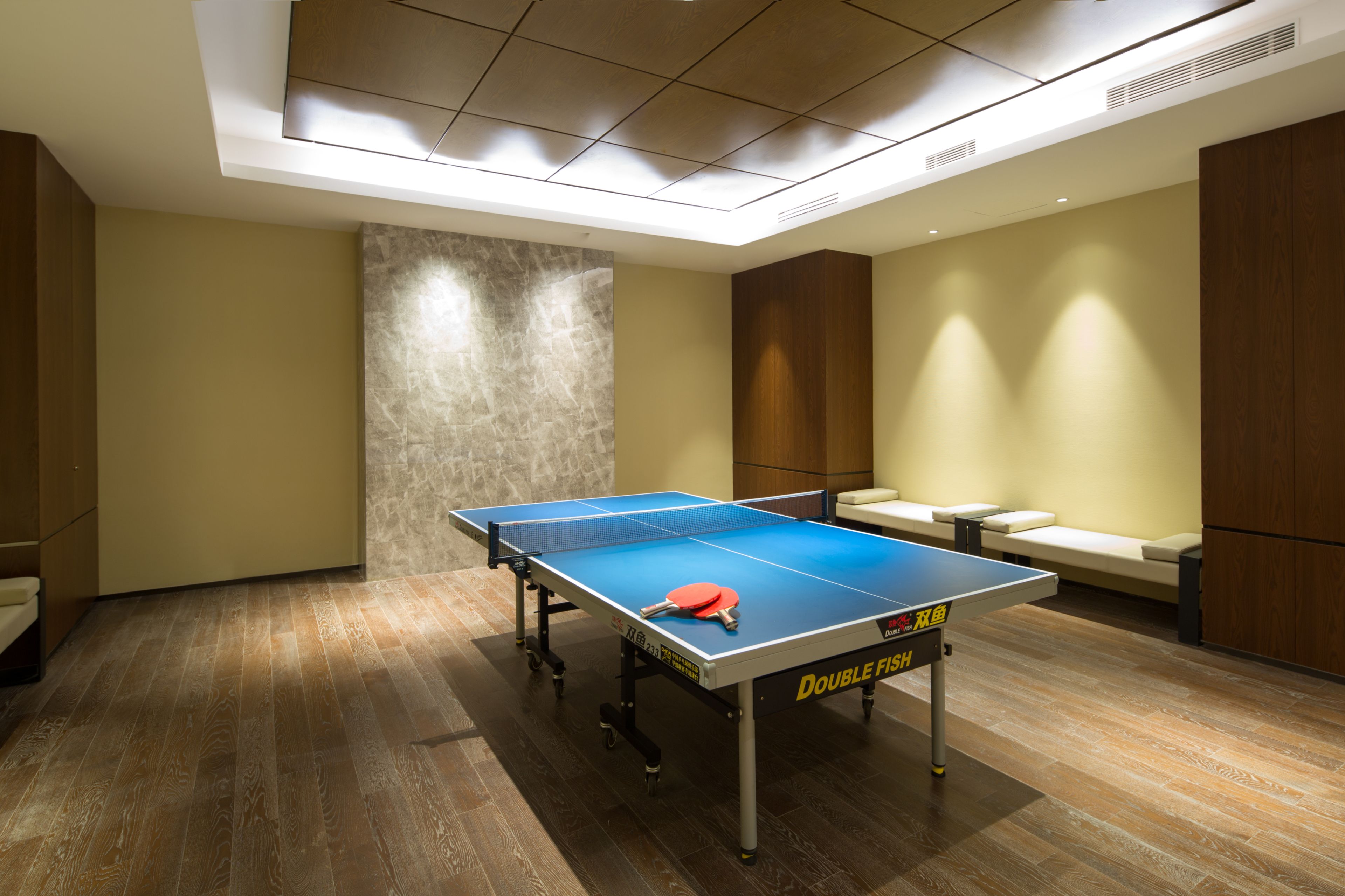 A ping pong table in a room with wooden flooring, beige walls, and a seating area with a sofa and chairs.