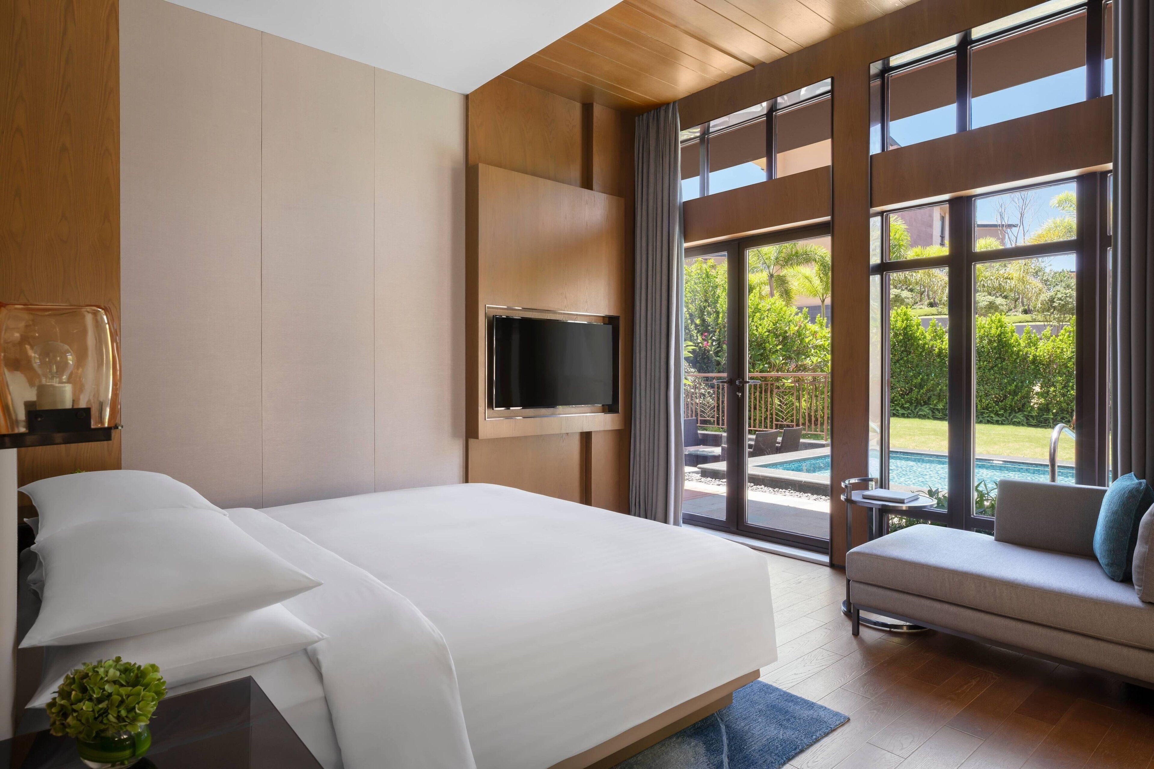 A modern hotel room with a large bed, a sofa, a TV mounted on the wall, and a view of a swimming pool and greenery.