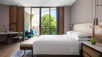 A modern hotel room with a large bed, a desk, and a balcony with a view of greenery.