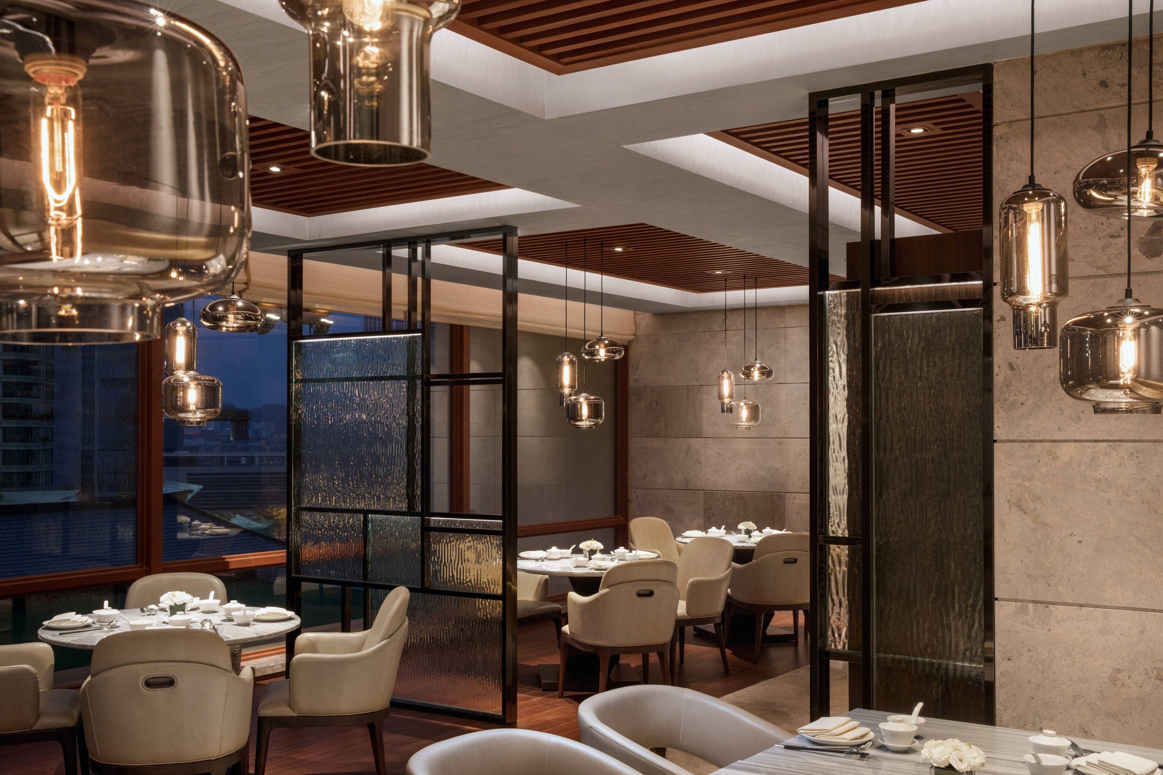 A modern restaurant with high ceilings, pendant lighting, and a view of the cityscape through large windows.