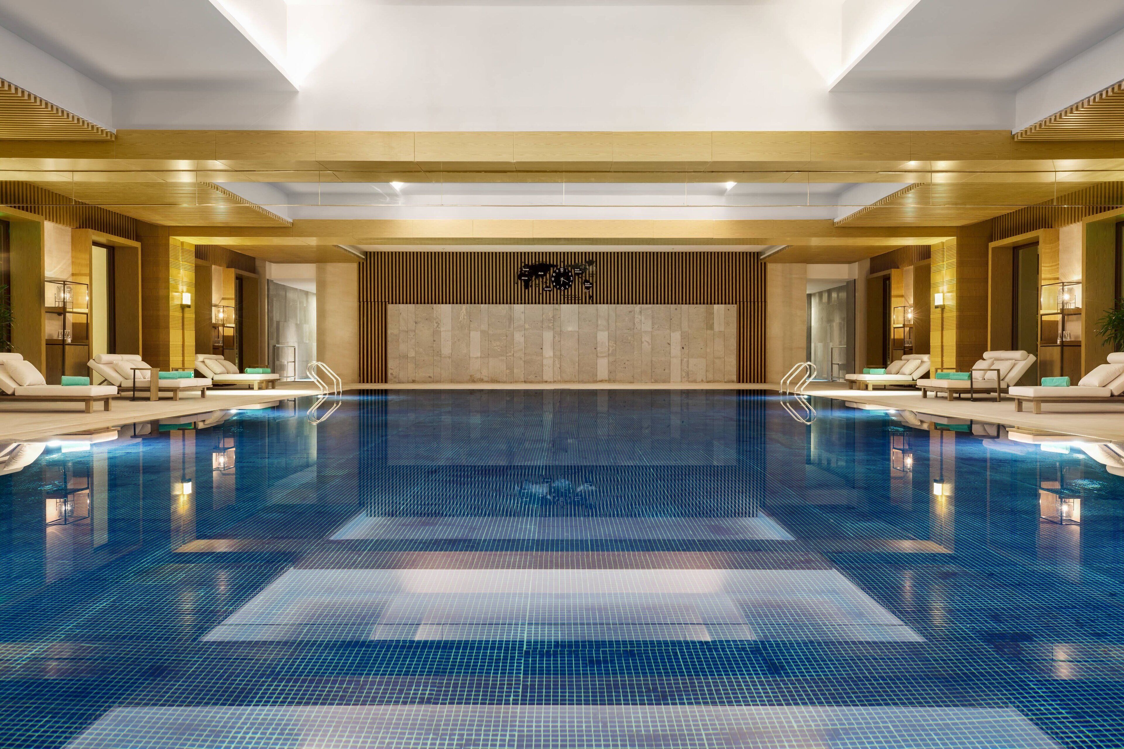 A large indoor swimming pool with lounge areas on either side, a wooden wall with a logo, and recessed lighting.
