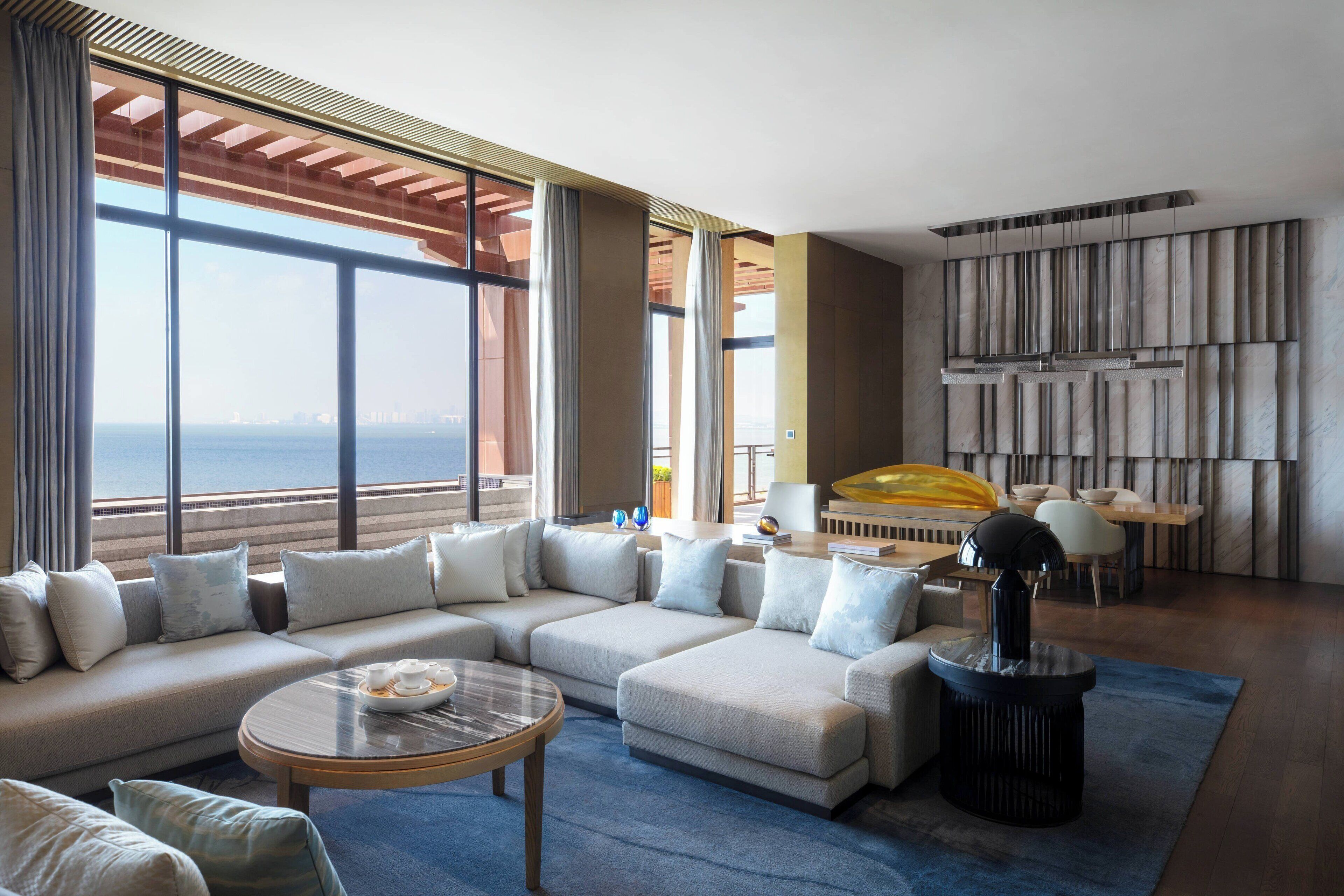 A modern living room with a large sectional sofa, a round coffee table, and a view of the sea through large windows.