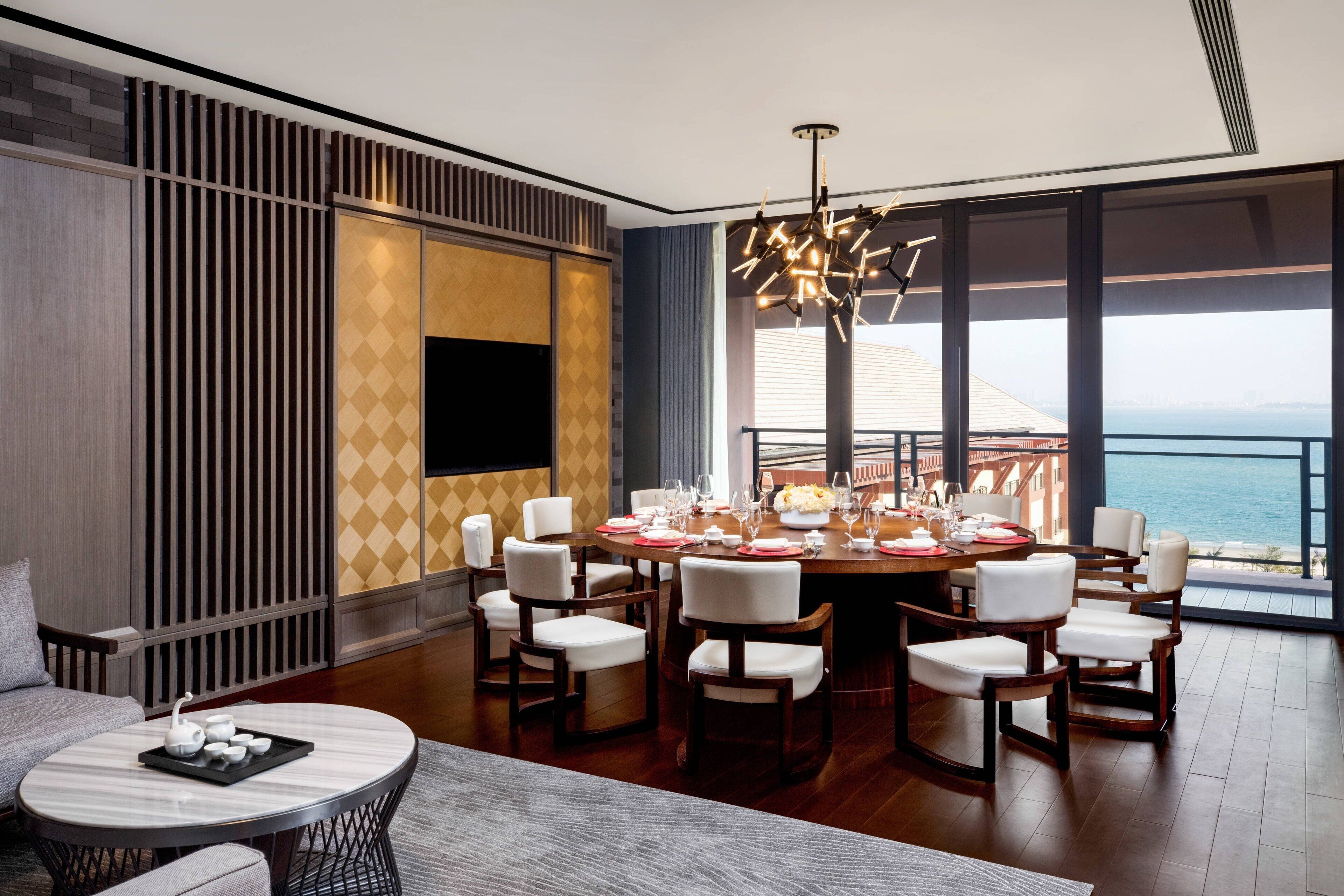 A dining area with a round table set for six, surrounded by chairs, with a view of the sea through large windows.